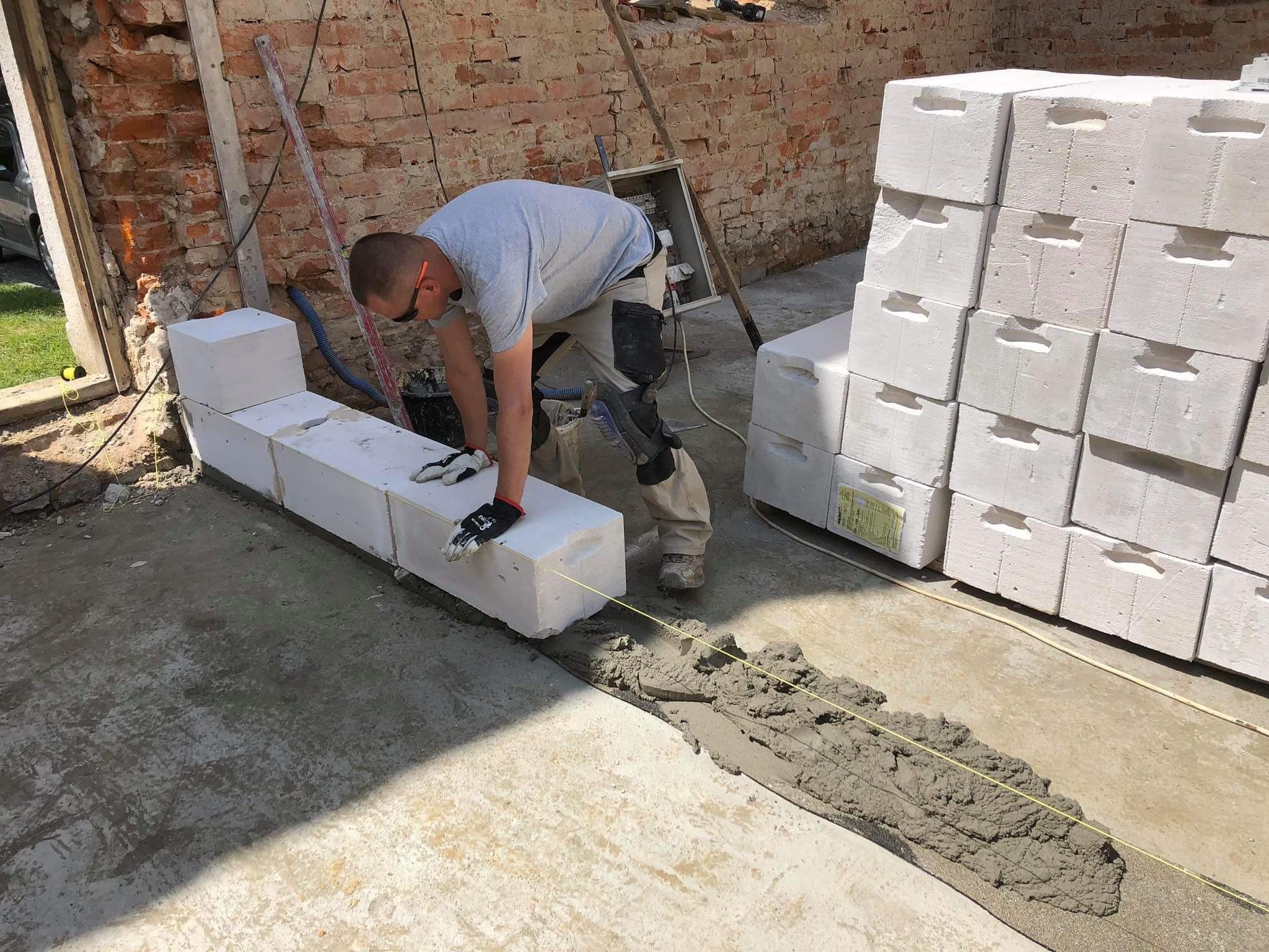Murarz układa bloczki z betonu komórkowego na fundamencie, widoczny sznurek traserski i zapas bloczków, ściana z czerwonej cegły w tle.