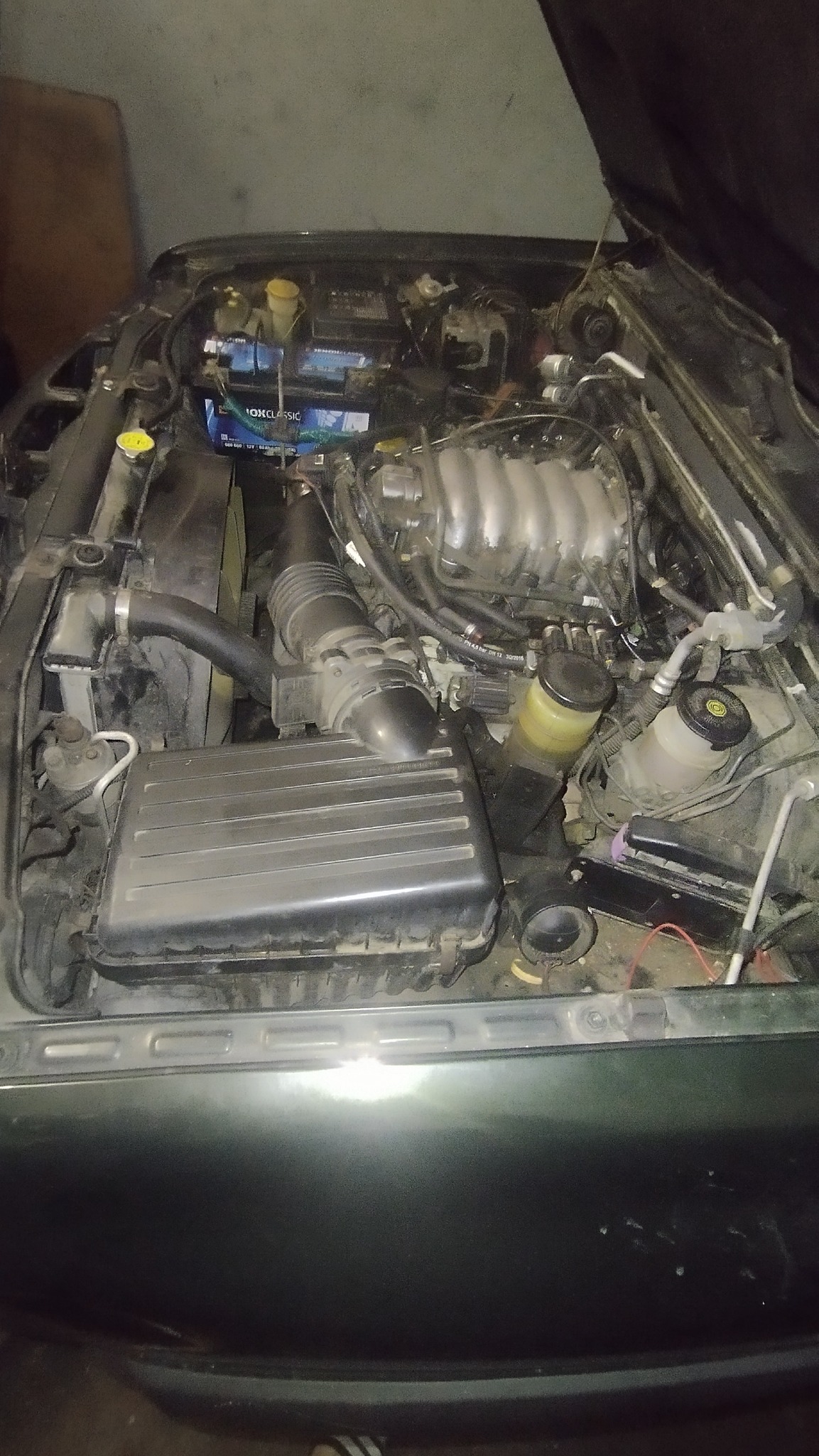 Otwarta maska samochodu z widocznym silnikiem, akumulatorem i innymi elementami. Widoczne przewody i płyny eksploatacyjne. Ujęcie z góry, ciemne oświetlenie.