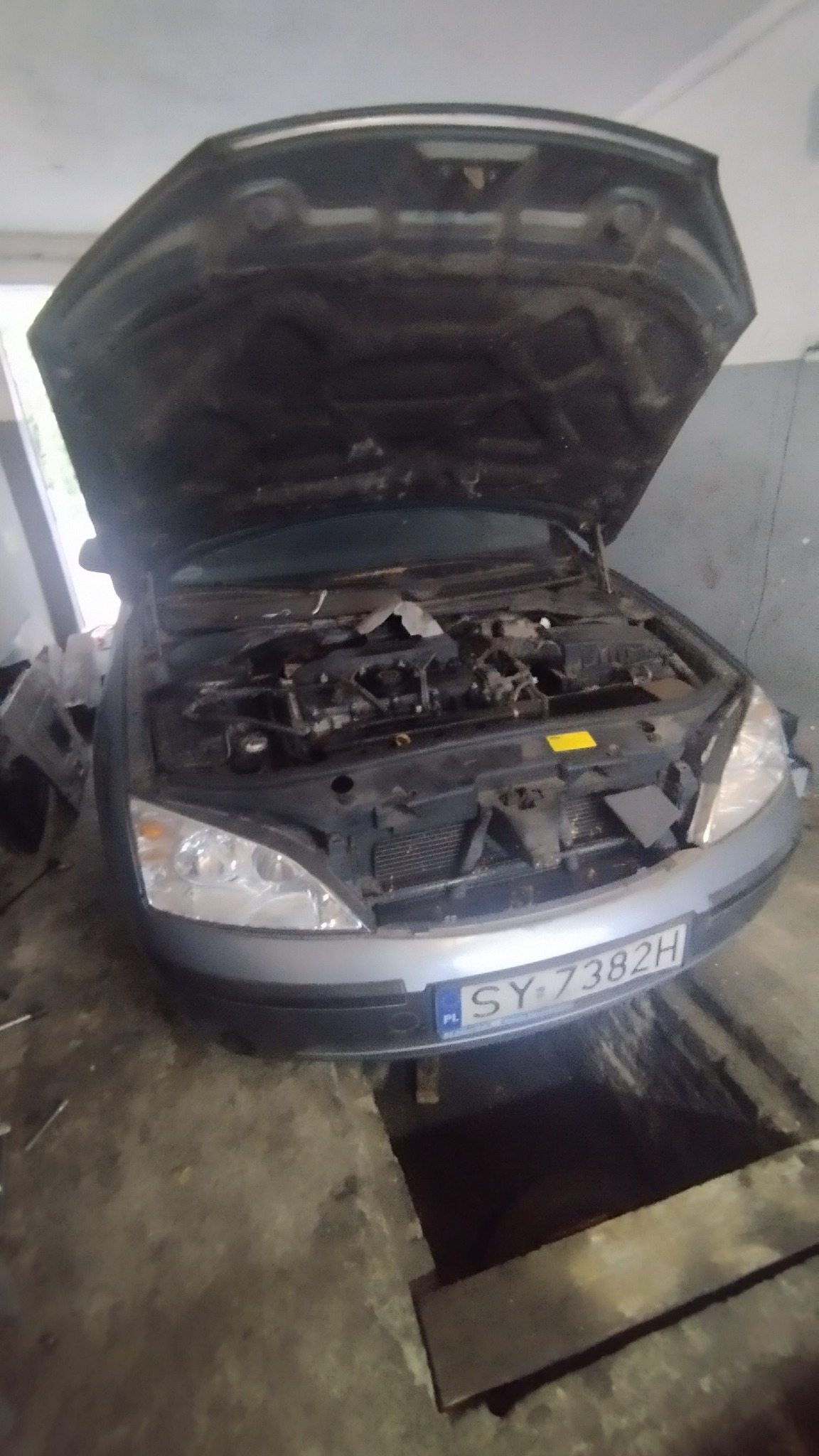 Szary samochód z otwartą maską w warsztacie, widoczny silnik i tablica rejestracyjna. Ujęcie z dołu, w tle ściana warsztatu i kanał samochodowy.