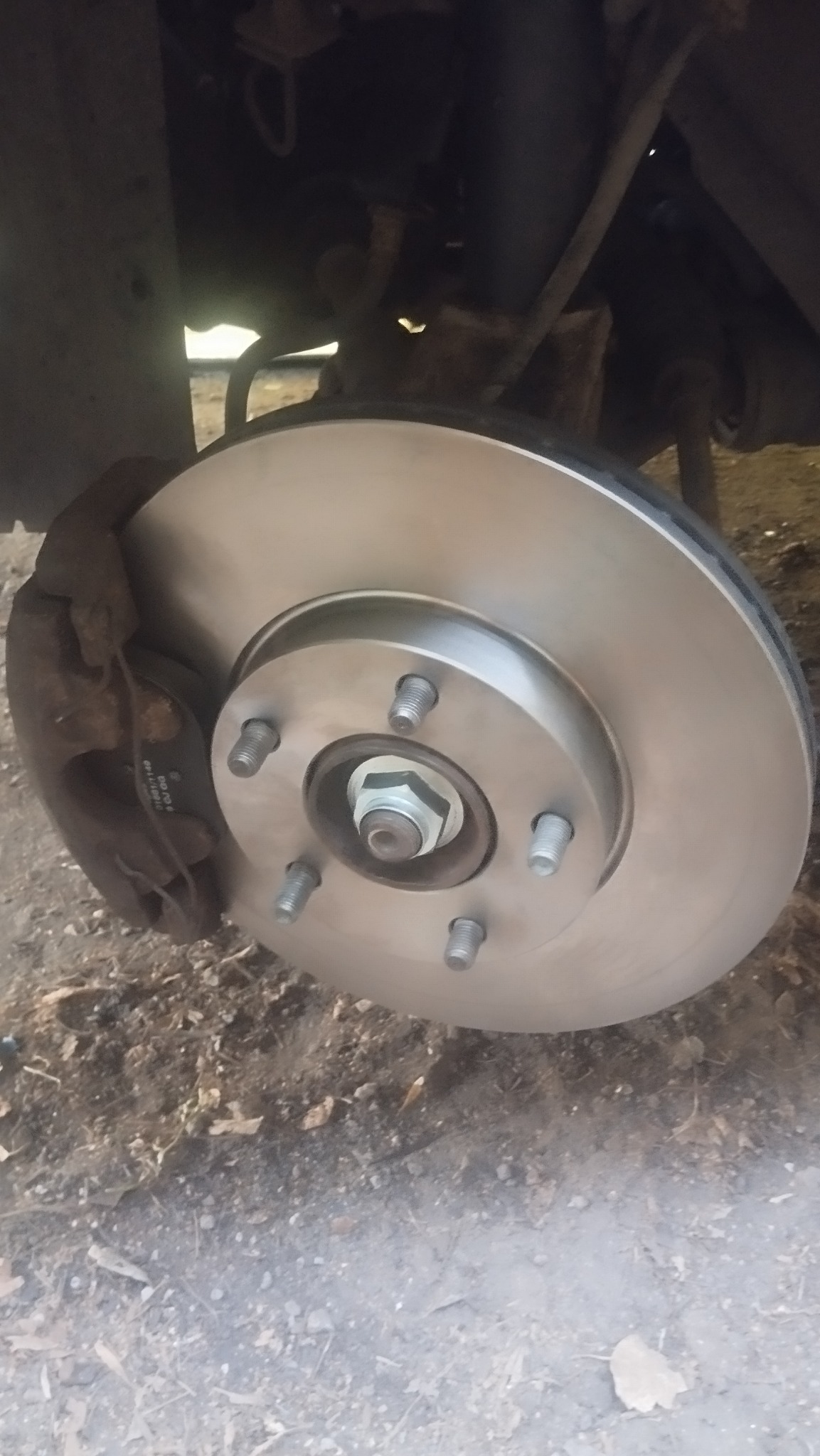 Nowa tarcza hamulcowa zamontowana w samochodzie, widoczne śruby mocujące i zacisk hamulcowy. Tło ziemiste, oświetlenie naturalne.