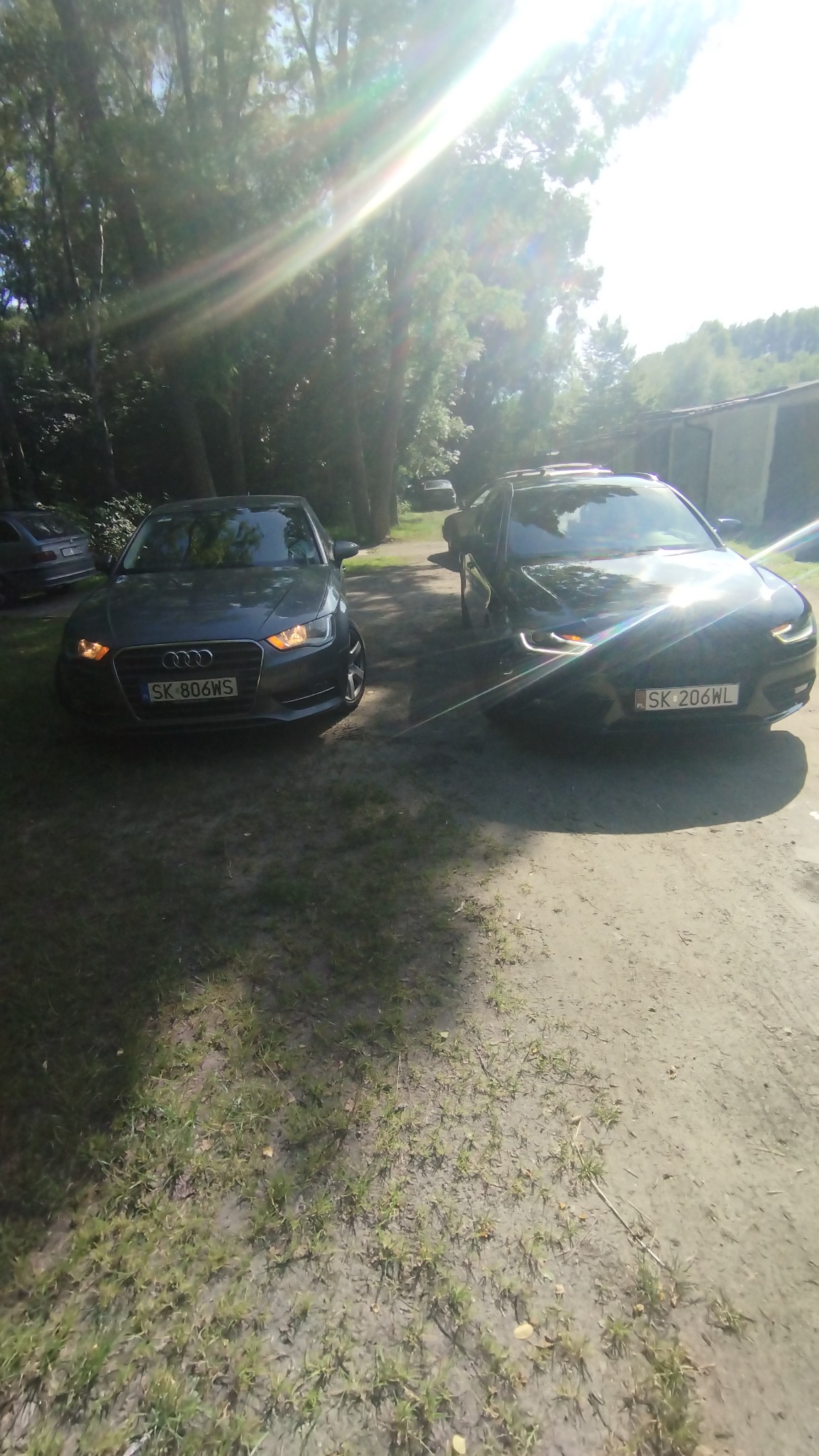 Dwa samochody Audi, szary A3 i czarny A4, zaparkowane na żwirowej drodze w słoneczny dzień. Widoczne tablice rejestracyjne i fragmenty otoczenia.