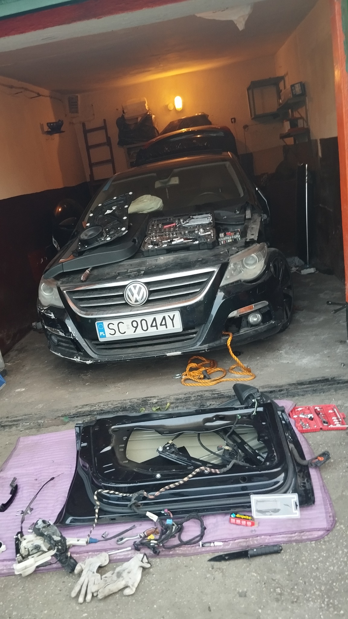 Czarny Volkswagen z otwartą maską i narzędziami w garażu. Zdemontowane drzwi leżą obok, ukazując wnętrze i elementy mechaniczne. Widoczne narzędzia i akcesoria.