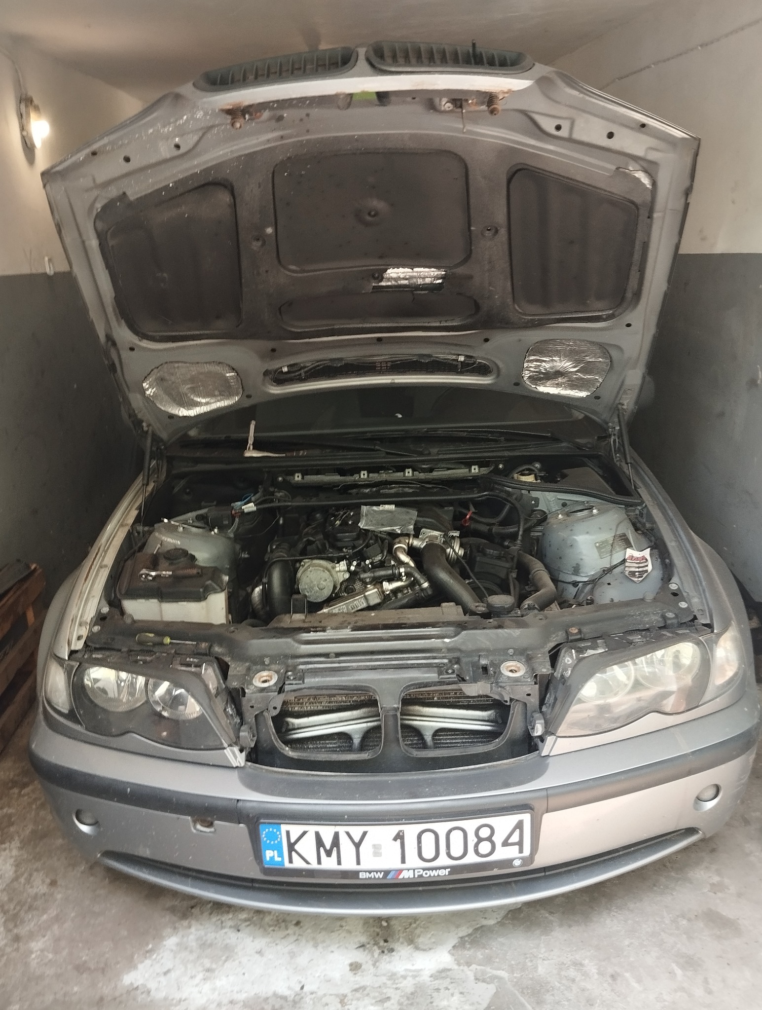 Srebrny samochód BMW z otwartą maską w garażu. Widoczny silnik, tablica rejestracyjna KMY 10084. Detale wnętrza komory silnika.