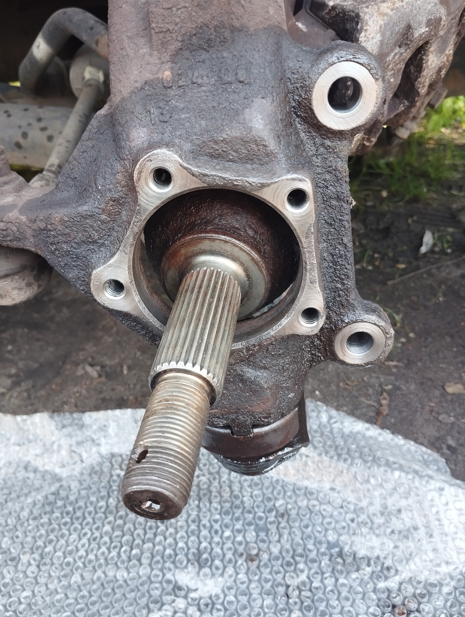 Piasta koła z łożyskiem i fragmentem osi, widoczne ślady smaru. Elementy zawieszenia pojazdu na tle podłoża z folii bąbelkowej.