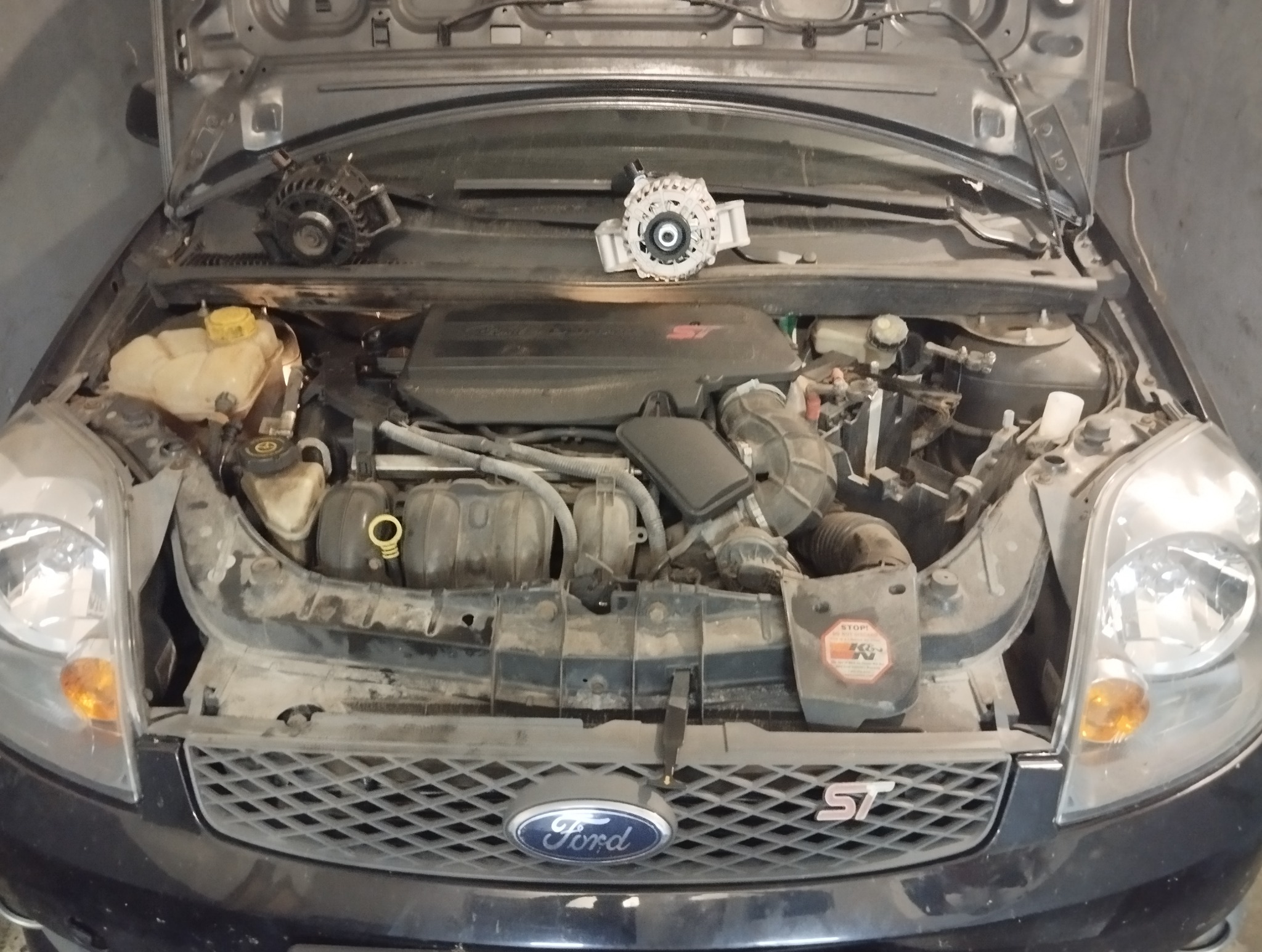 Otwarta maska czarnego Forda Fiesta ST z widocznym silnikiem i elementami osprzętu, w tym alternatorem leżącym na podszybiu. Widoczne logo Forda na grillu.