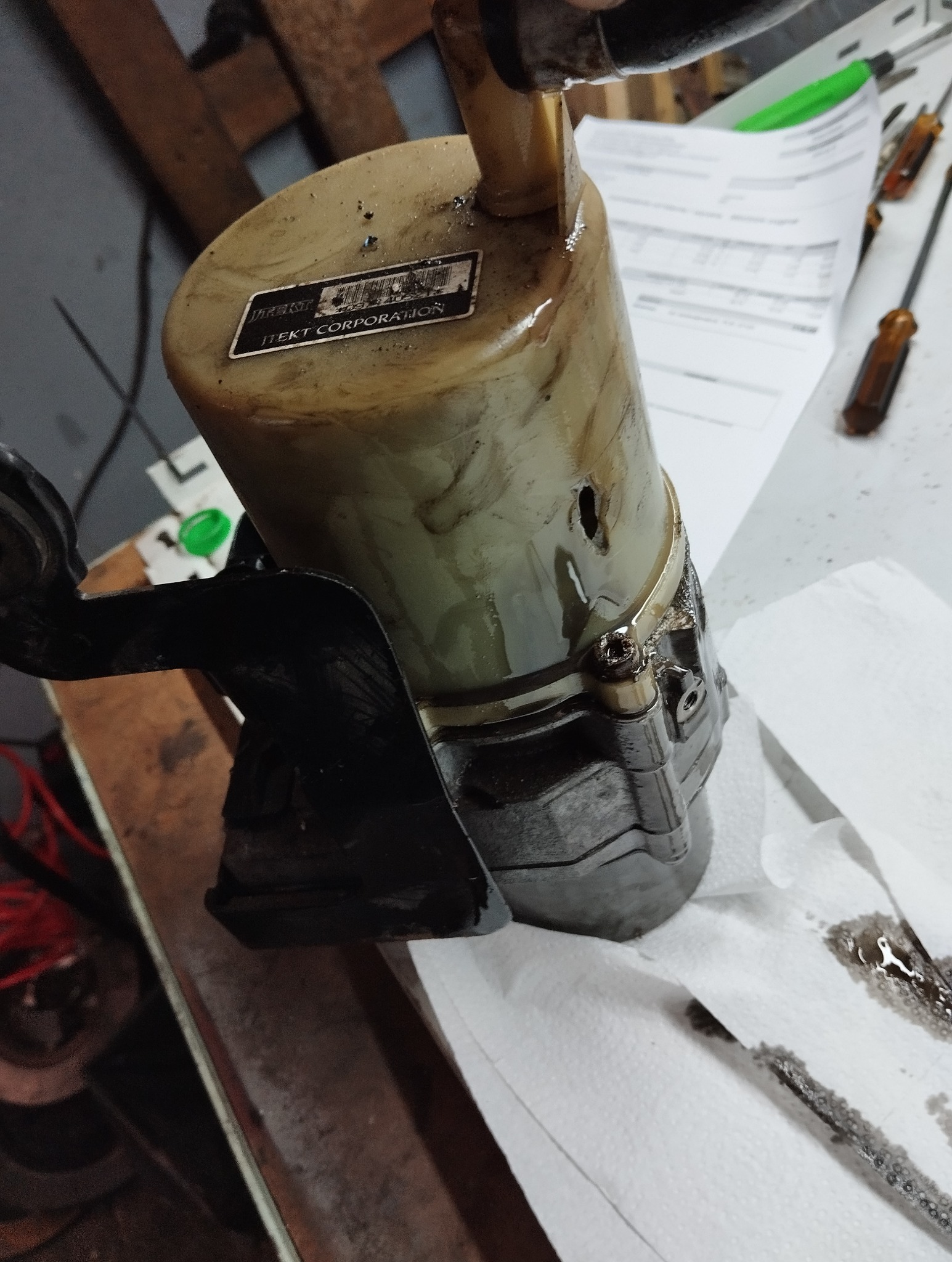 Brudny zbiornik płynu wspomagania układu kierowniczego ITEKT CORPORATION, z widocznymi śladami wycieków, na warsztatowym stole. W tle narzędzia i dokumentacja.