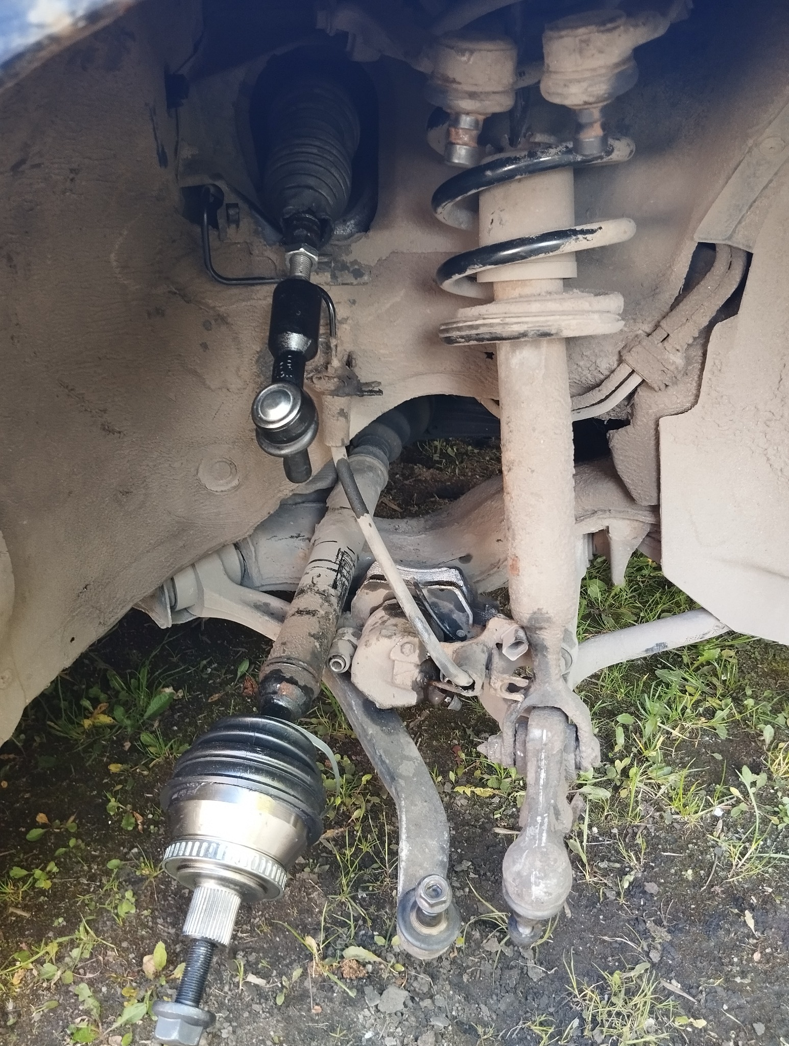 Elementy zawieszenia samochodu w trakcie naprawy, widoczne sprężyny, amortyzatory i przegub. Prace wykonywane na zewnątrz, na trawie.