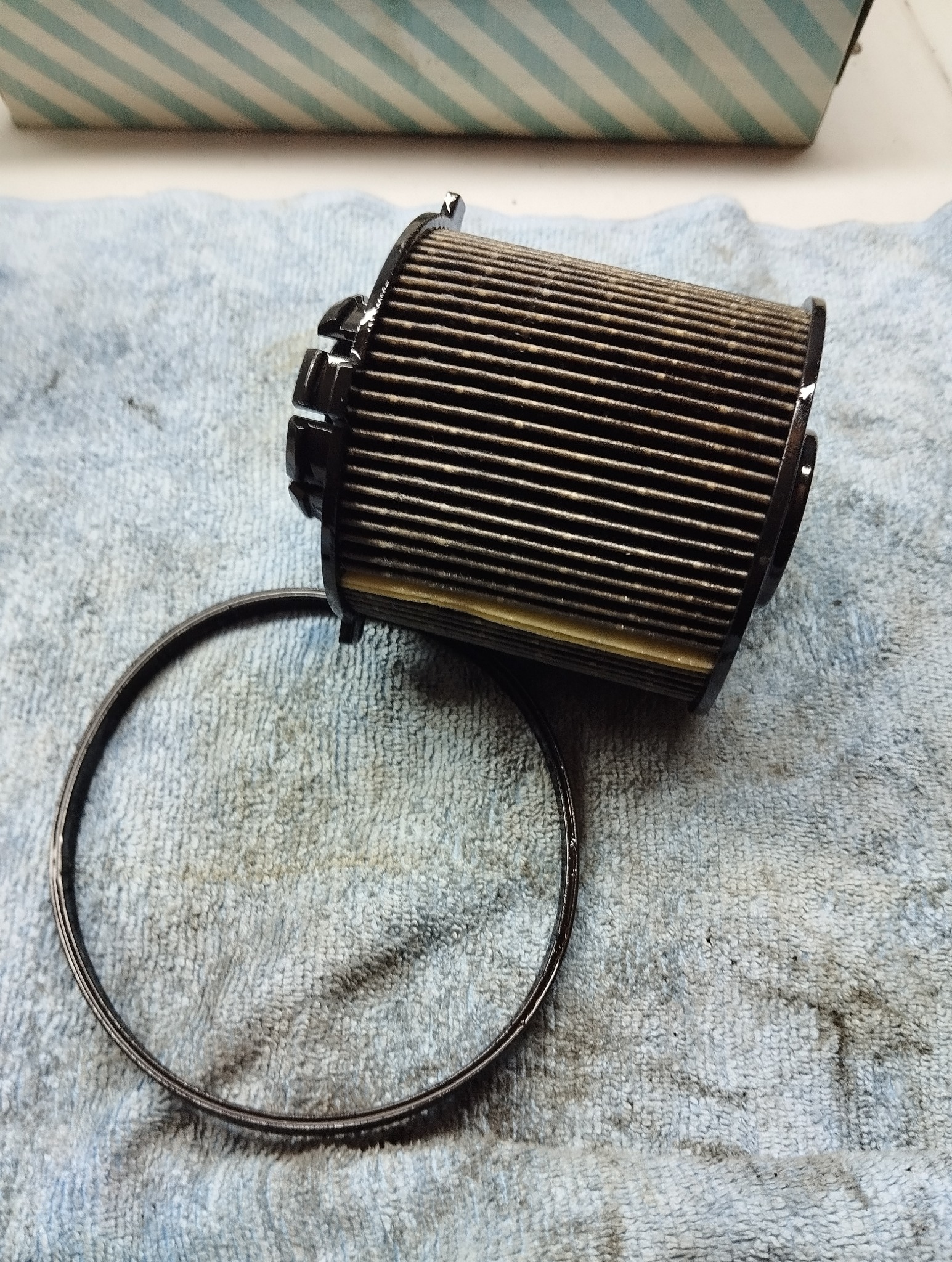 Używany, cylindryczny filtr oleju samochodowego z gumową uszczelką na tle szarej, teksturowanej ściereczki. Widoczne ślady zużycia.