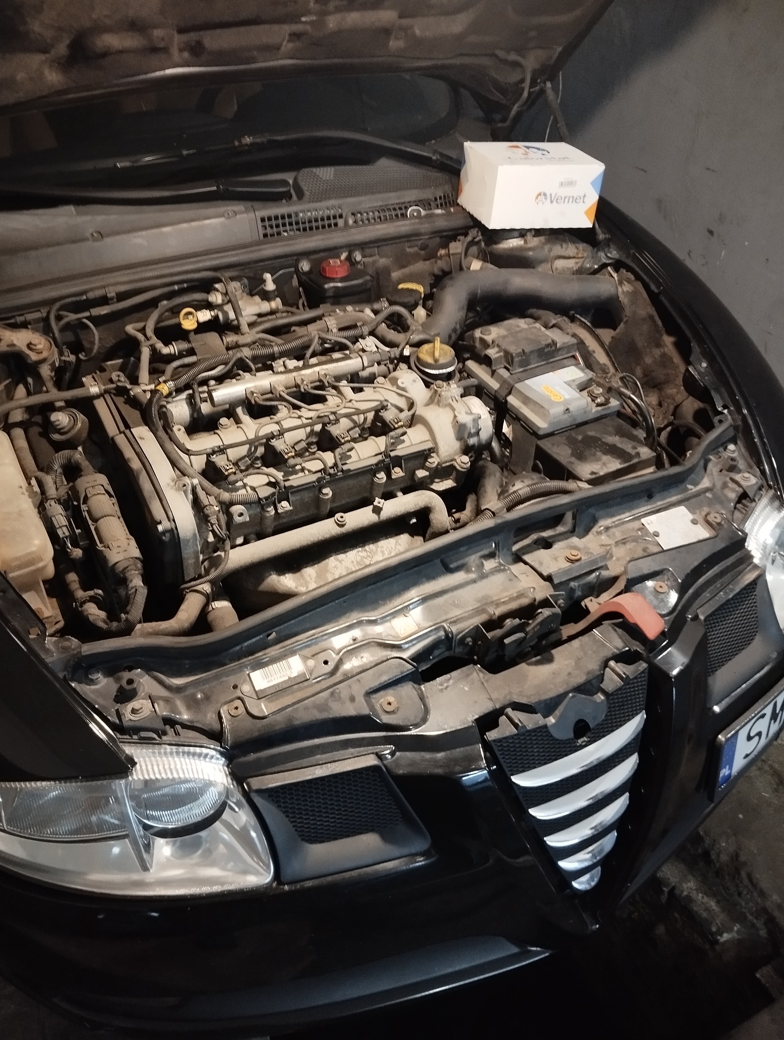 Otwarta maska czarnego samochodu Alfa Romeo z widocznym silnikiem i pudełkiem części zamiennych marki Vernet na górze. Widoczne elementy silnika i akumulator.