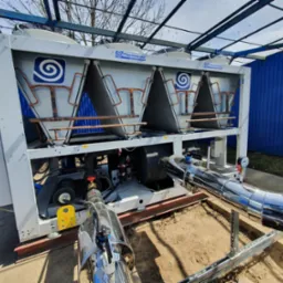 Zewnętrzna instalacja chłodnicza Thermocold z wentylatorami, rurami miedzianymi i izolowanymi rurami stalowymi, widok z dołu na tle niebieskiej ściany i drzew