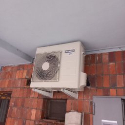 COLD MAN Maciej Wysokiński - Zewnętrzna jednostka klimatyzacji Hitachi zamontowana na ceglanej ścianie budynku, widoczne metalowe wsporniki, fragment drzwi i ogłoszenie na nich.