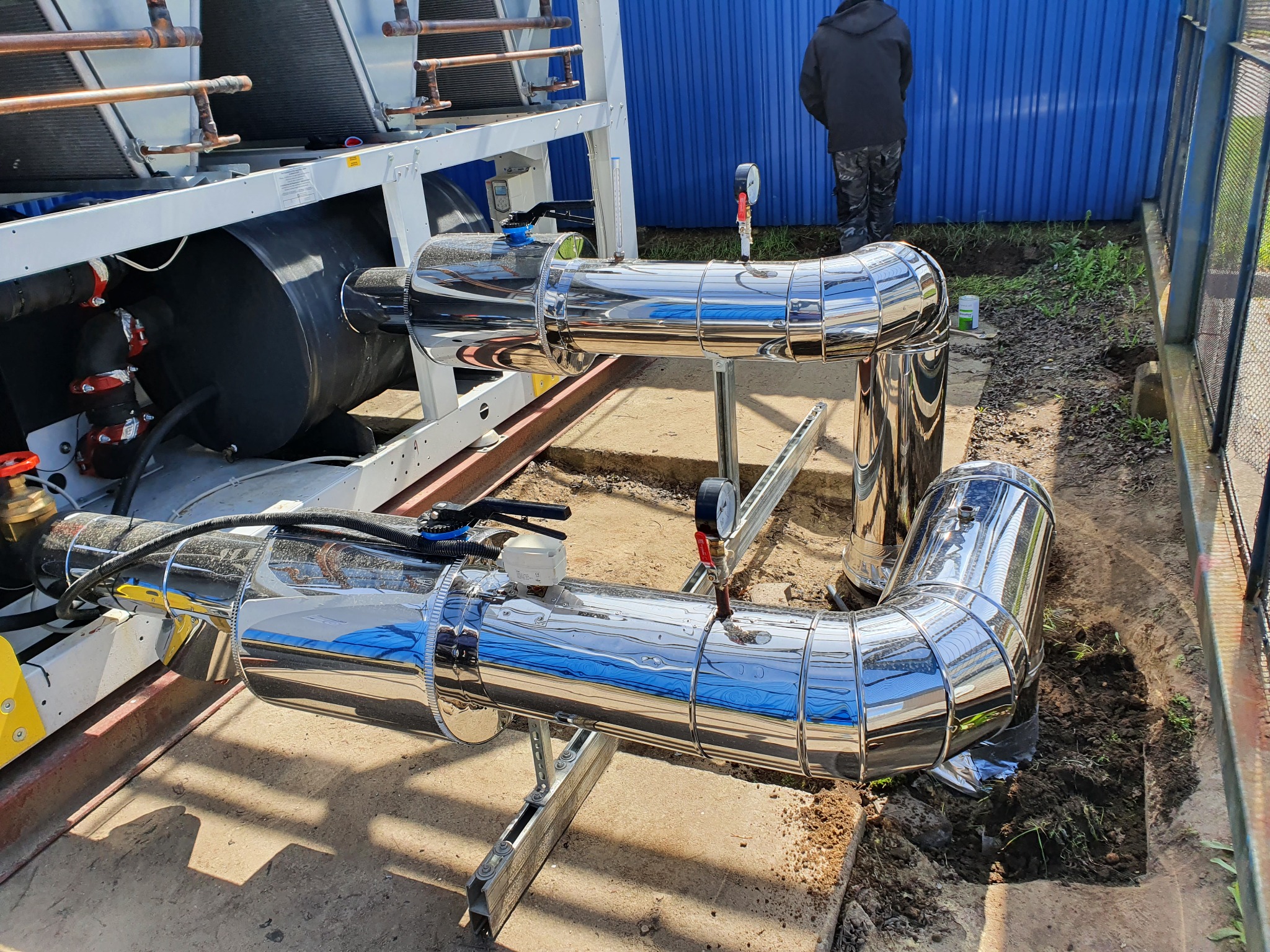 Błyszczące, chromowane rury instalacji hydraulicznej z manometrami, zamontowane na zewnątrz budynku, widoczny fragment urządzenia i osoba w oddali przy niebieskiej ścianie.