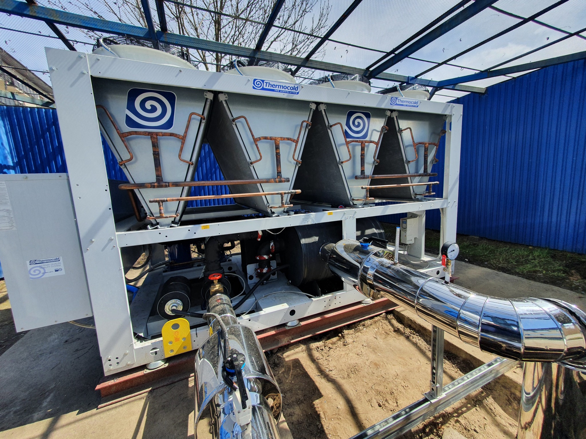 Zewnętrzna instalacja chłodnicza Thermocold z wentylatorami, rurami miedzianymi i izolowanymi rurami stalowymi, widok z dołu na tle niebieskiej ściany i drzew