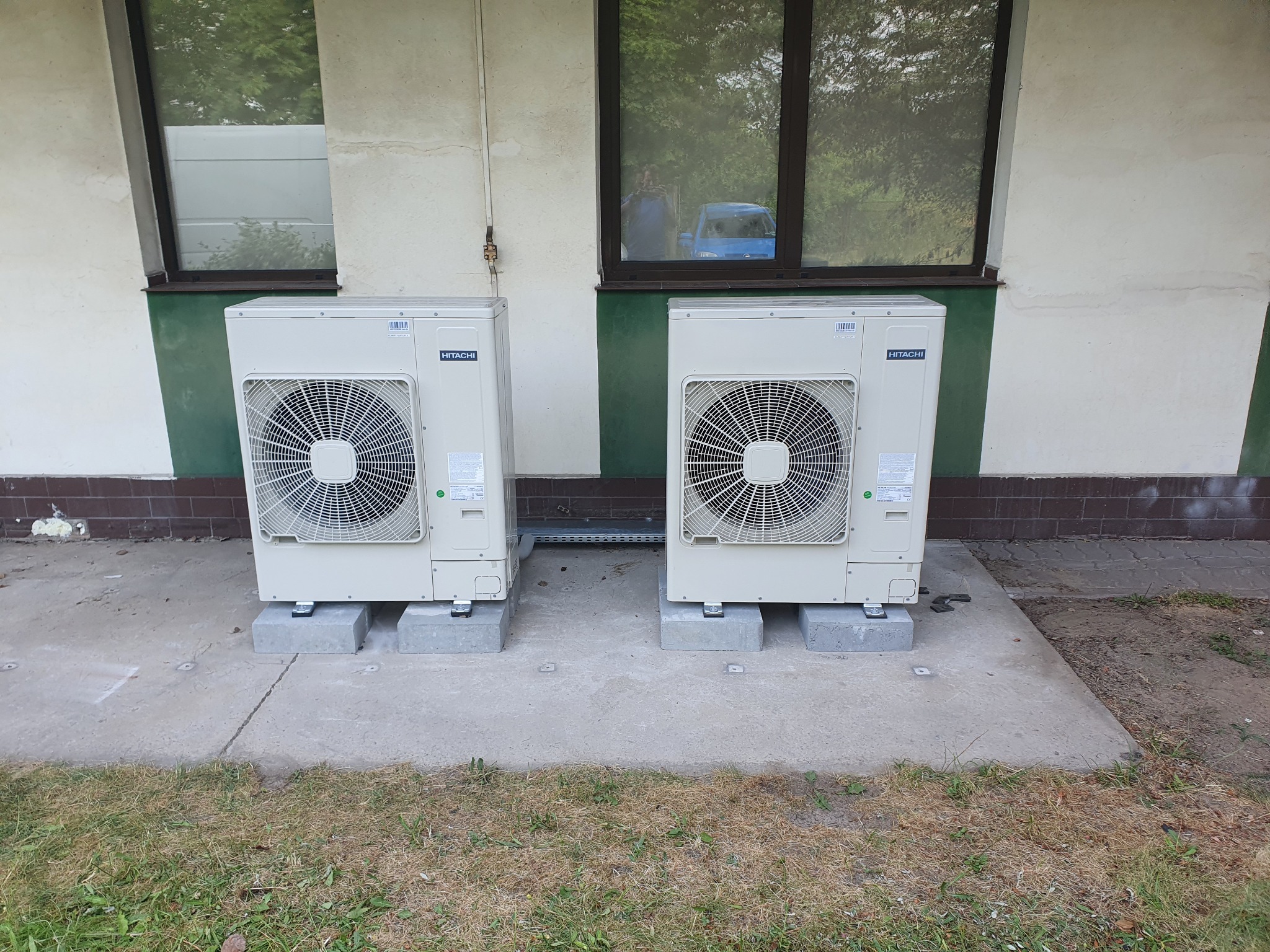 Dwa zewnętrzne agregaty klimatyzacji Hitachi, zamontowane na betonowych podstawach przed budynkiem z zielonymi akcentami, widoczne przez okno odbicie osoby robiącej zdjęcie i niebieskiego samochodu.