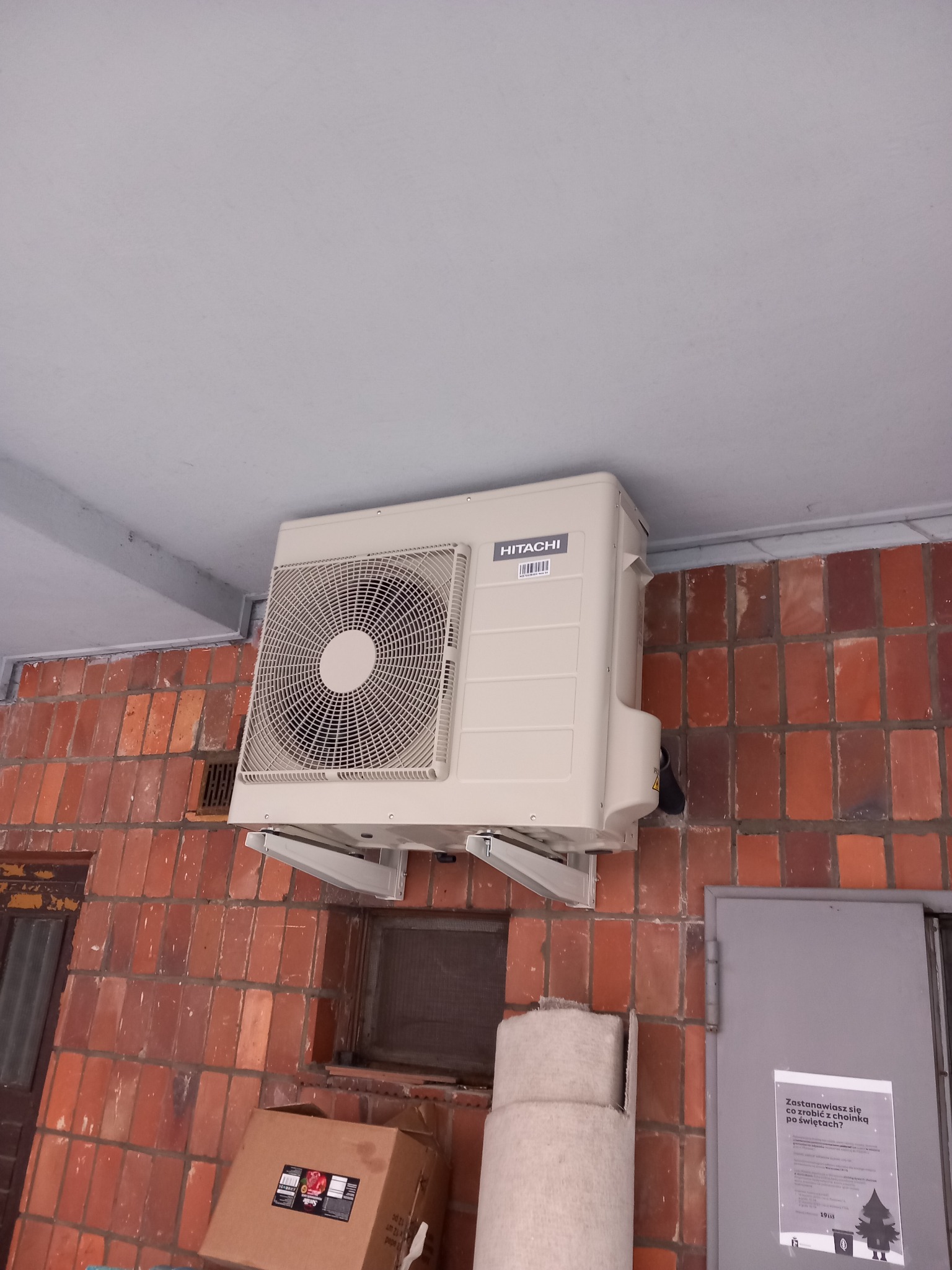 Zewnętrzna jednostka klimatyzacji Hitachi zamontowana na ceglanej ścianie budynku, widoczne metalowe wsporniki, fragment drzwi i ogłoszenie na nich.