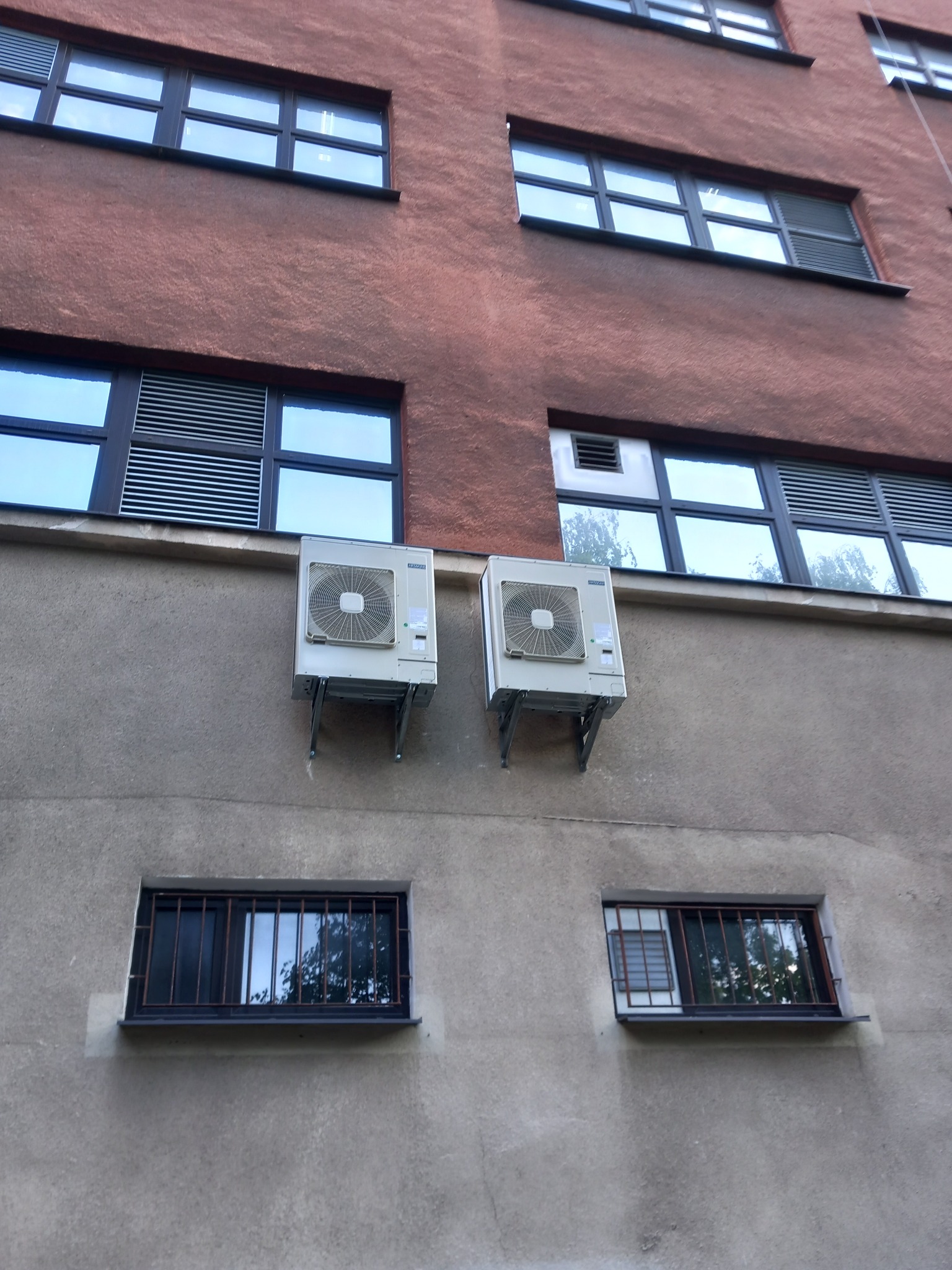 Dwa klimatyzatory zamontowane na szarym murze budynku, widoczne okna z kratami i roletami, elewacja budynku w kolorze ceglastym.