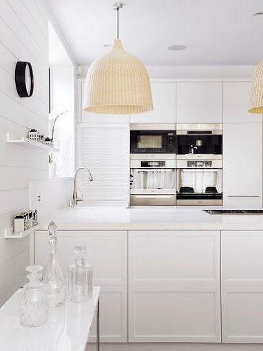 Jasna, minimalistyczna kuchnia z białymi frontami, oświetleniem w stylu boho i szklanymi naczyniami dekoracyjnymi na blacie.