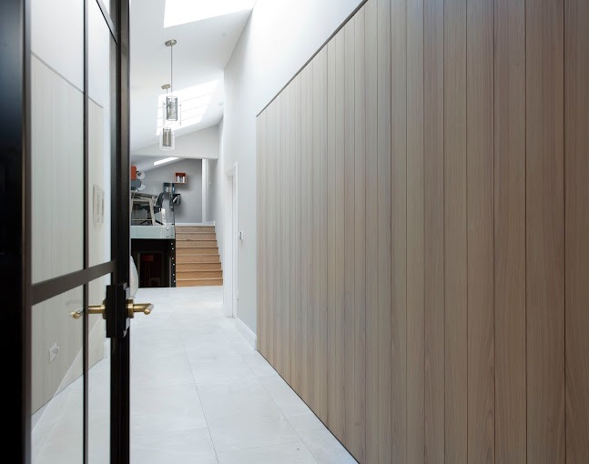 Wnętrze korytarza z okładziną ścienną z jasnego drewna, widok przez otwarte, czarne, przeszklone drzwi, w tle schody i wiszące lampy.