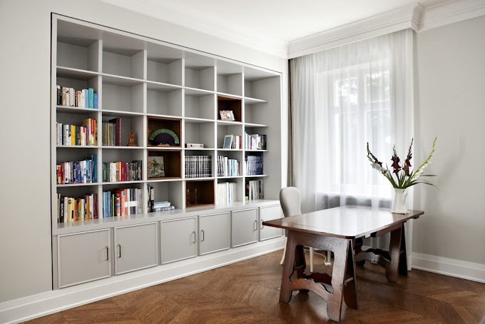 Jasnoszara zabudowa biblioteczna z szafkami dolnymi i otwartymi półkami, wypełniona książkami i dekoracjami, ustawiona w pokoju z parkietem w jodełkę i drewnianym biurkiem pod oknem z firanką.