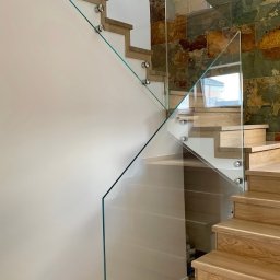 Sun-Glass Michał Gregotowicz - Nowoczesne schody z drewnianymi stopniami zabezpieczone szklaną balustradą mocowaną punktowo, na ścianie dekoracyjna okładzina z kamienia naturalnego.
