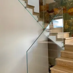 Nowoczesne schody z drewnianymi stopniami zabezpieczone szklaną balustradą mocowaną punktowo, na ścianie dekoracyjna okładzina z kamienia naturalnego.