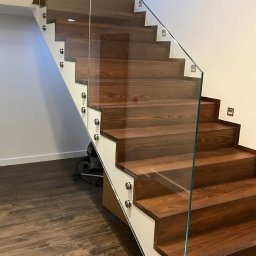 Sun-Glass Michał Gregotowicz - Nowoczesne schody z drewnianymi stopniami i szklaną balustradą mocowaną na metalowych uchwytach, oświetlenie w stopniach, widok z dołu na górę.