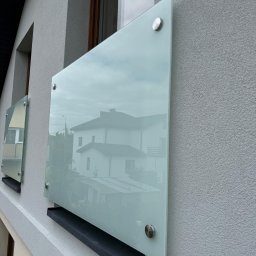 Sun-Glass Michał Gregotowicz - Szklane panele przymocowane do ściany budynku za pomocą metalowych śrub, odbijające otoczenie, w tym budynek mieszkalny i drzewa.