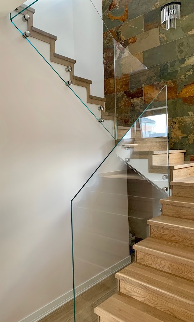 Nowoczesne schody z drewnianymi stopniami zabezpieczone szklaną balustradą mocowaną punktowo, na ścianie dekoracyjna okładzina z kamienia naturalnego.