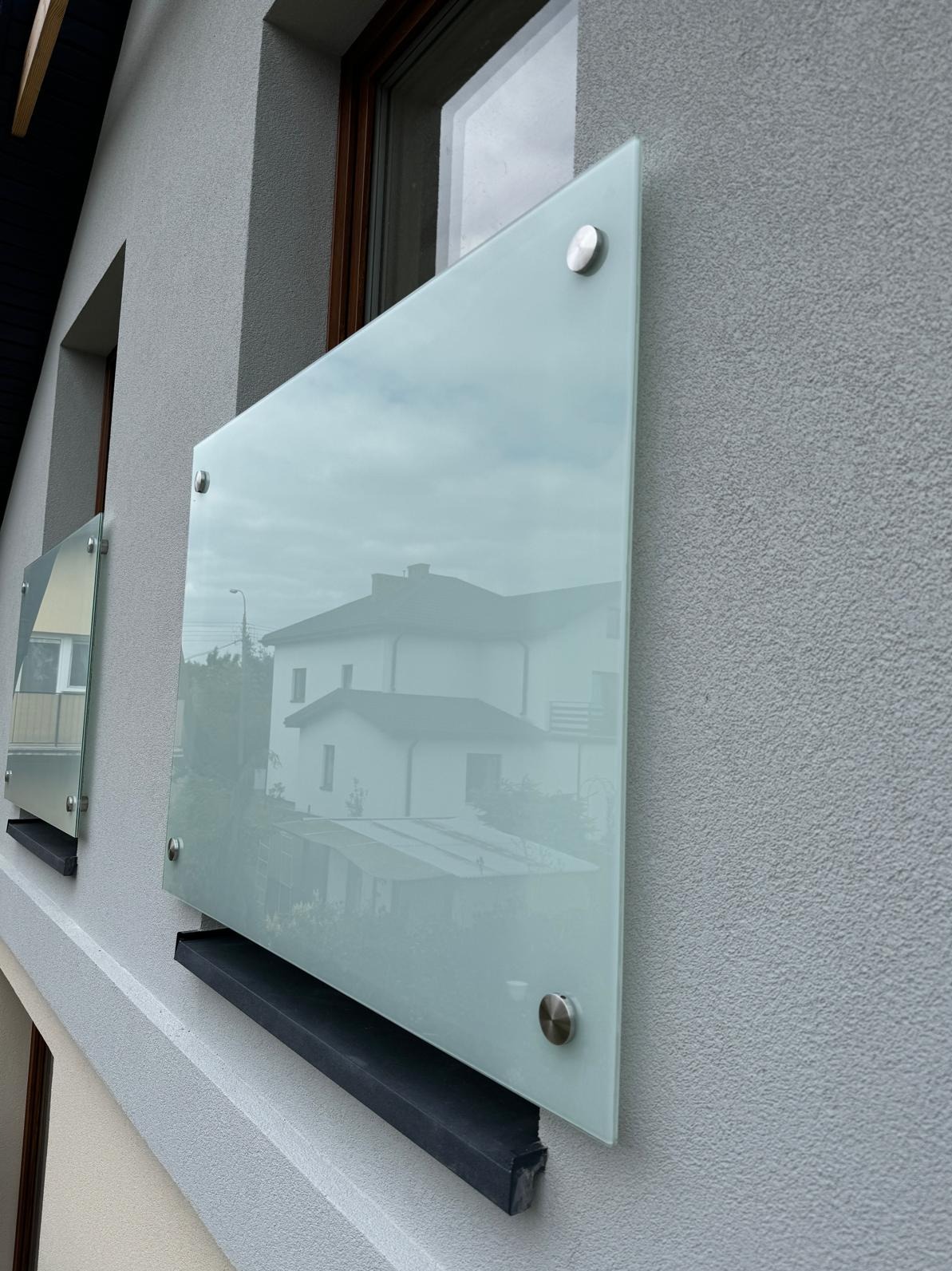 Szklane panele przymocowane do ściany budynku za pomocą metalowych śrub, odbijające otoczenie, w tym budynek mieszkalny i drzewa.