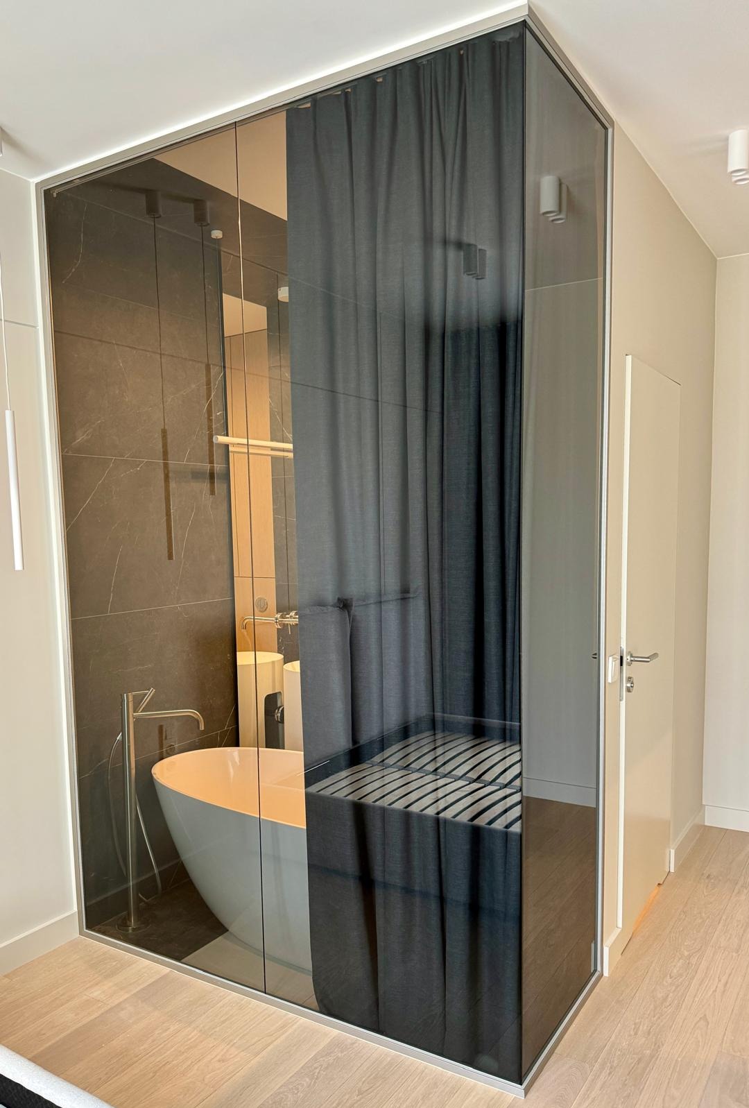 Szklana kabina prysznicowa typu walk-in z grafitowymi zasłonami, widoczna wanna wolnostojąca i armatura w tle, jasna drewniana podłoga.