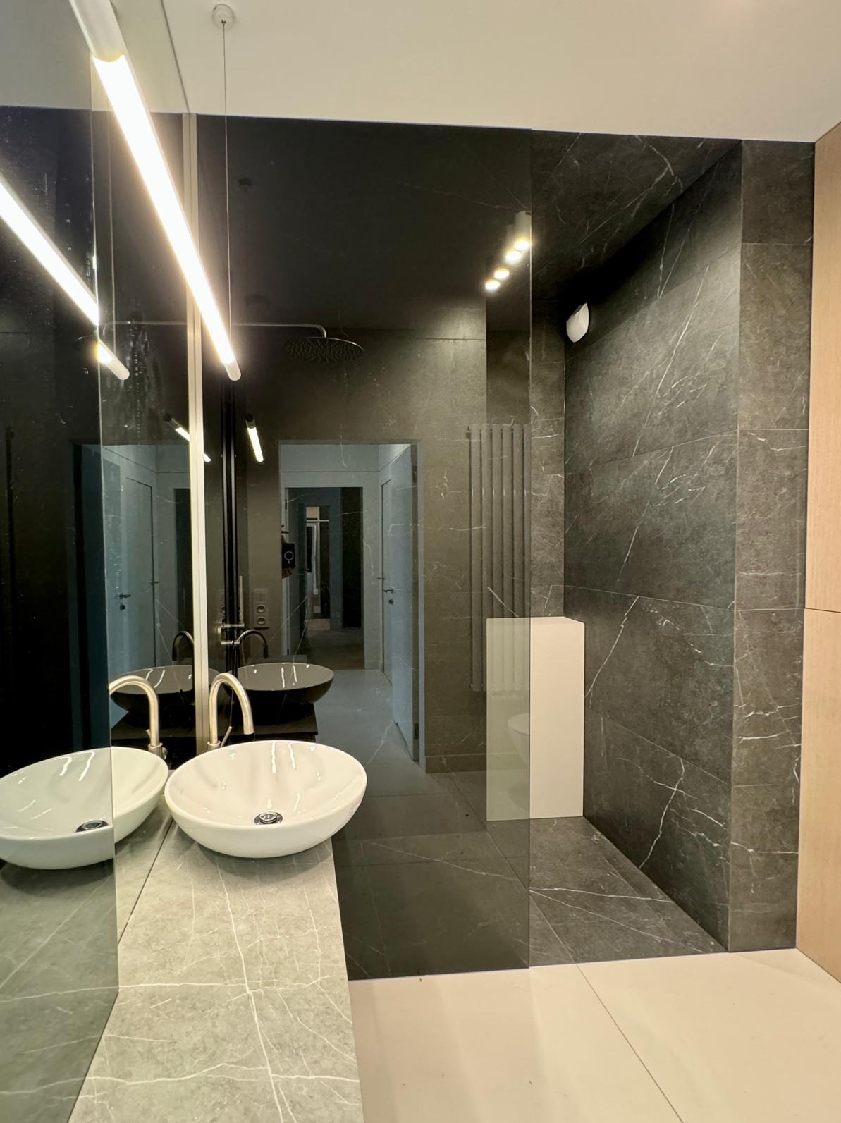 Nowoczesna łazienka z umywalkami nablatowymi, szklaną ścianą prysznicową i grafitową okładziną ścienną z marmurowym wzorem.