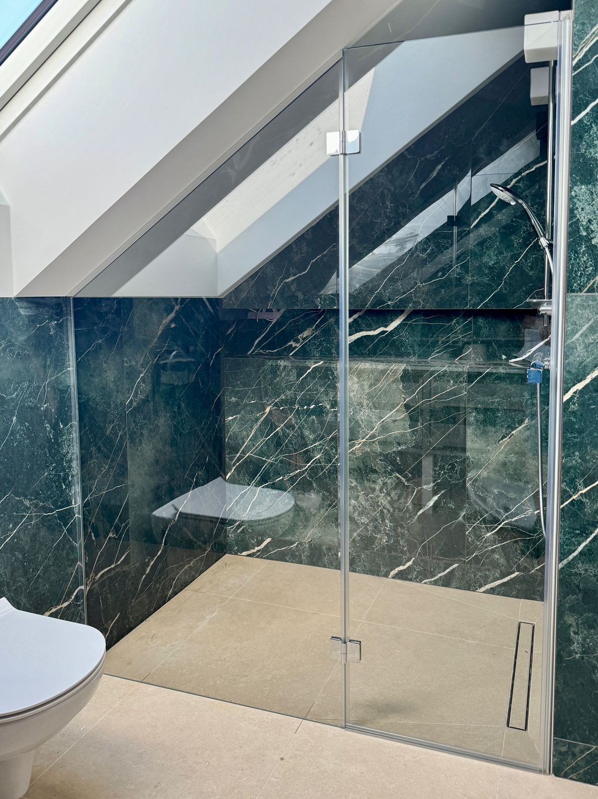Kabina prysznicowa ze szklaną ścianką w łazience z zielonym marmurem, widoczny fragment toalety i skośny sufit.