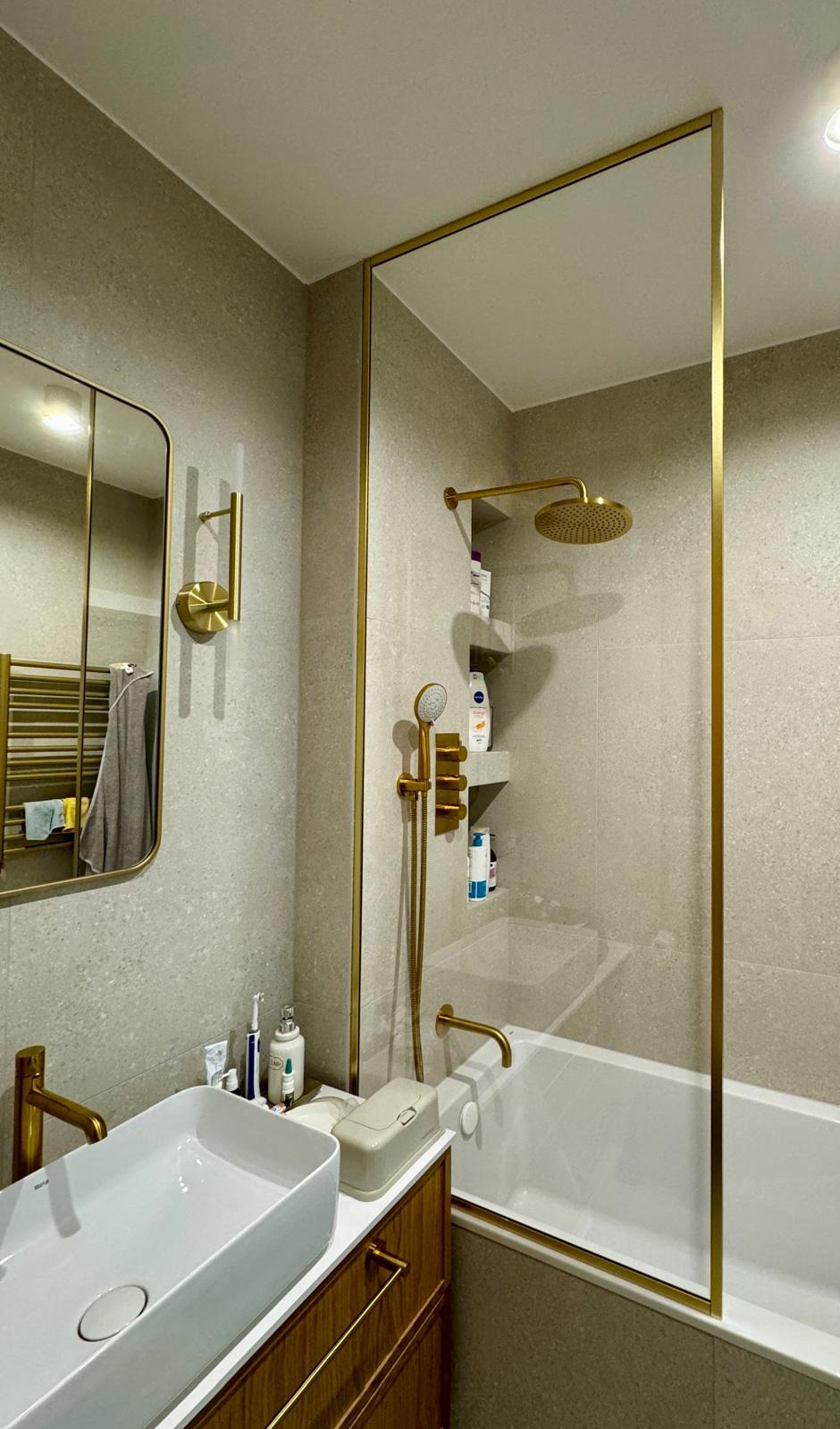 Nowoczesna łazienka z kabiną prysznicową oddzieloną szklaną ścianką w złotej ramie, armatura w kolorze złota, umywalka nablatowa i lustro z podświetleniem.