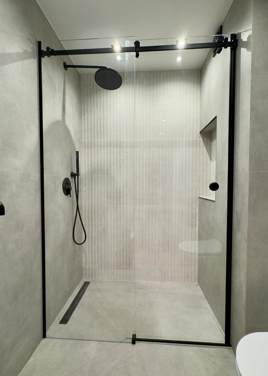 Nowoczesna kabina prysznicowa z przesuwnymi drzwiami ze szkła, czarne profile, deszczownica i odpływ liniowy w szarej łazience.