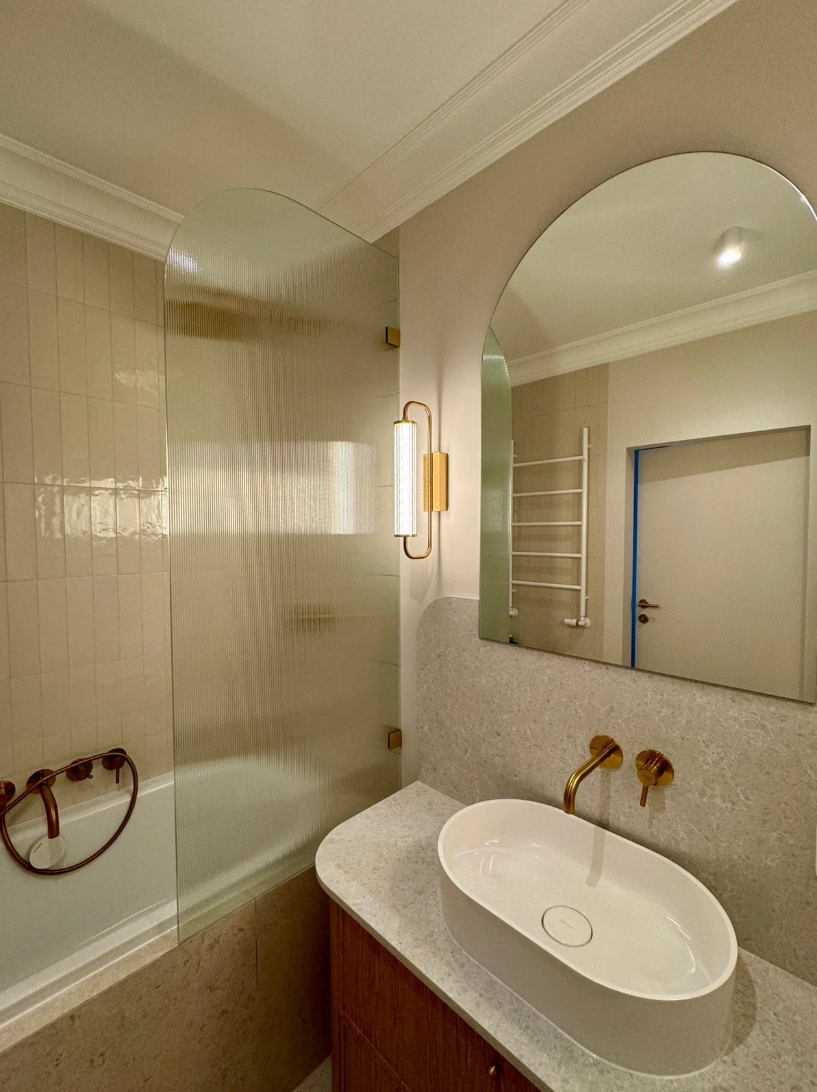 Nowoczesna łazienka z kabiną prysznicową z ryflowanego szkła, owalną umywalką nablatową i złotą armaturą, oświetlona lampą ścienną w stylu art deco.