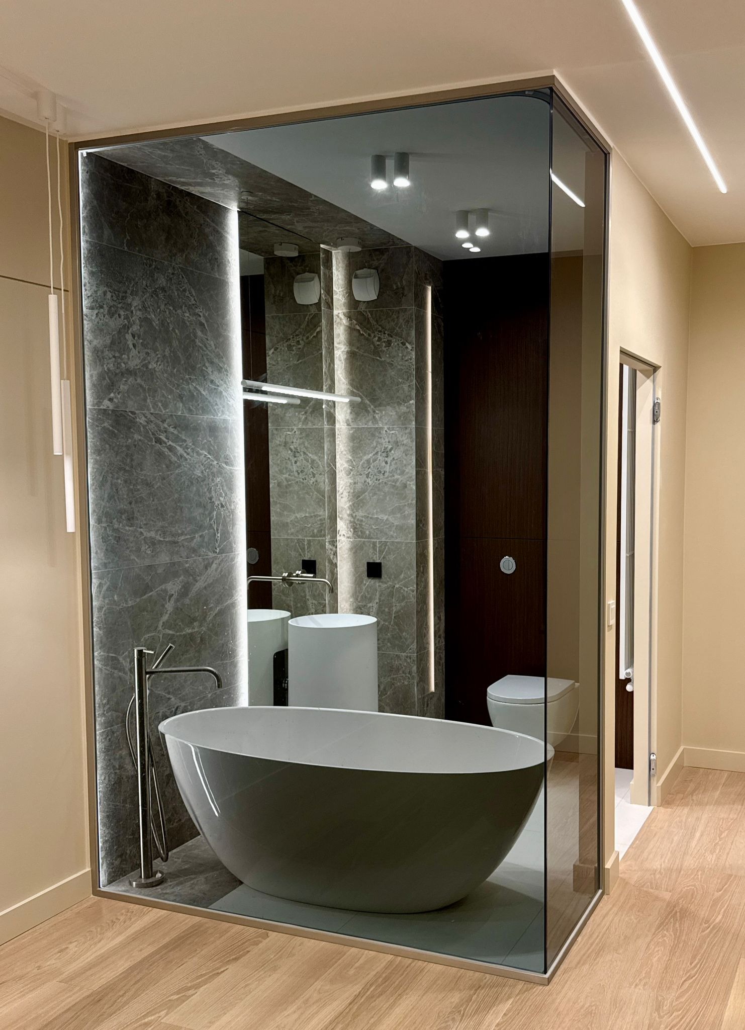 Nowoczesna łazienka z wolnostojącą wanną, szklaną ścianą działową i marmurową okładziną, oświetlona listwami LED.