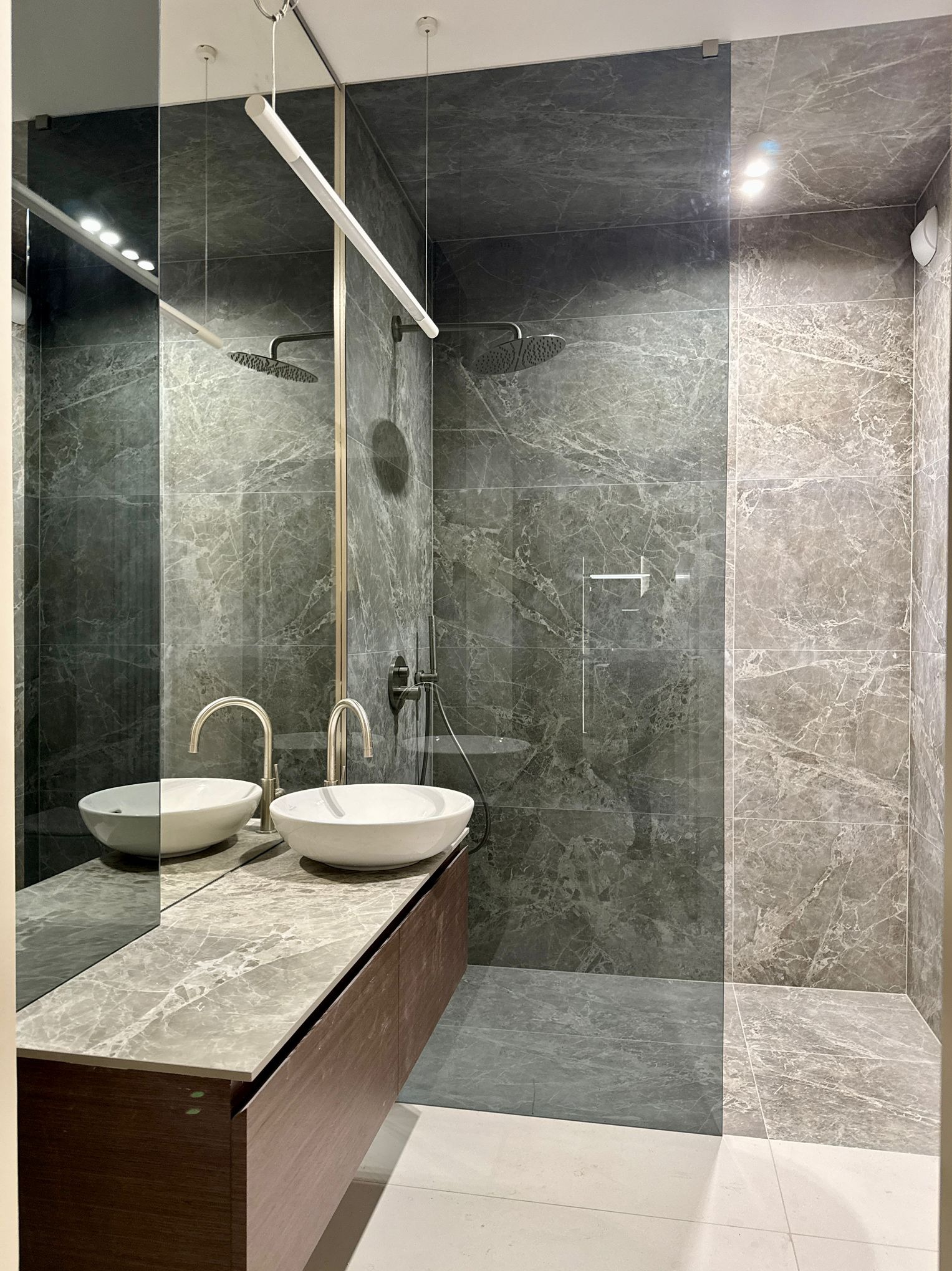 Nowoczesna łazienka z kabiną prysznicową oddzieloną szklaną ścianką, umywalkami nablatowymi i marmurowymi płytkami w odcieniach szarości.