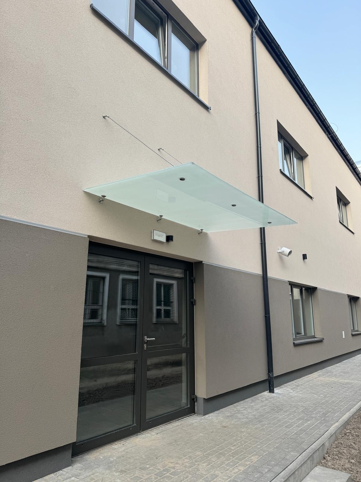 Szklany daszek nad wejściem do budynku, wsparty na metalowych uchwytach i linkach, z widocznymi drzwiami z przeszkleniami i elewacją w odcieniach beżu i szarości.