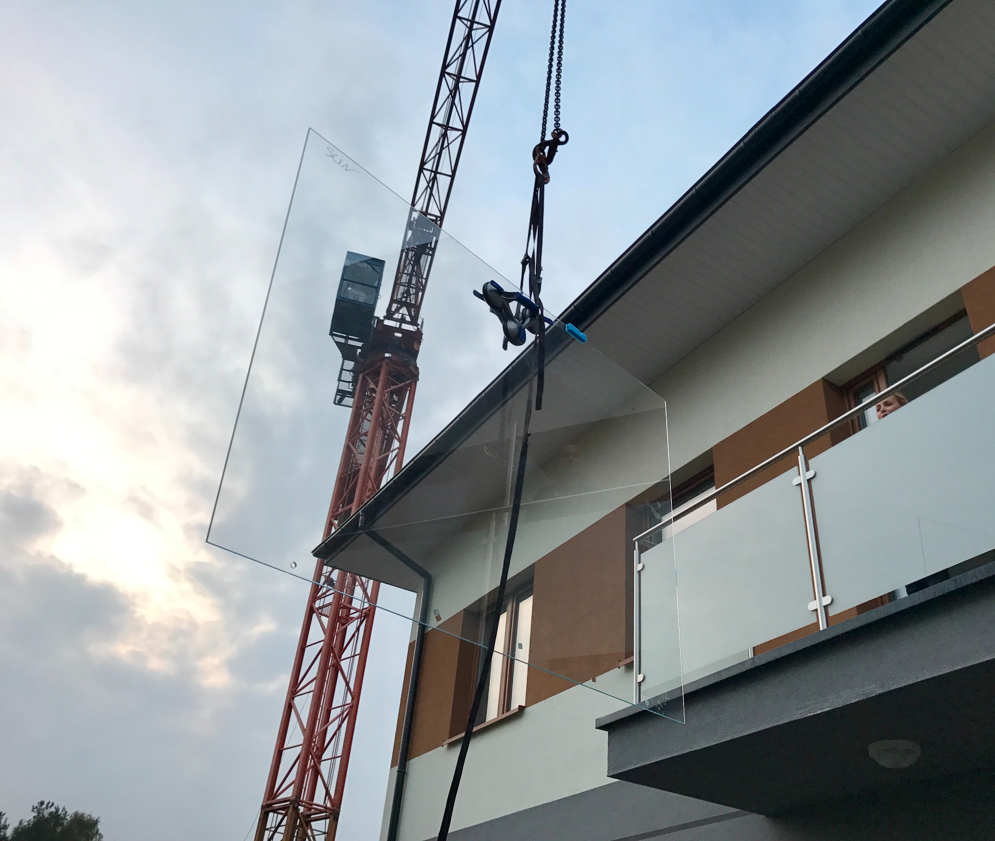Montaż tafli szkła na balustradzie balkonowej przy użyciu dźwigu, widoczna kobieta w oknie.