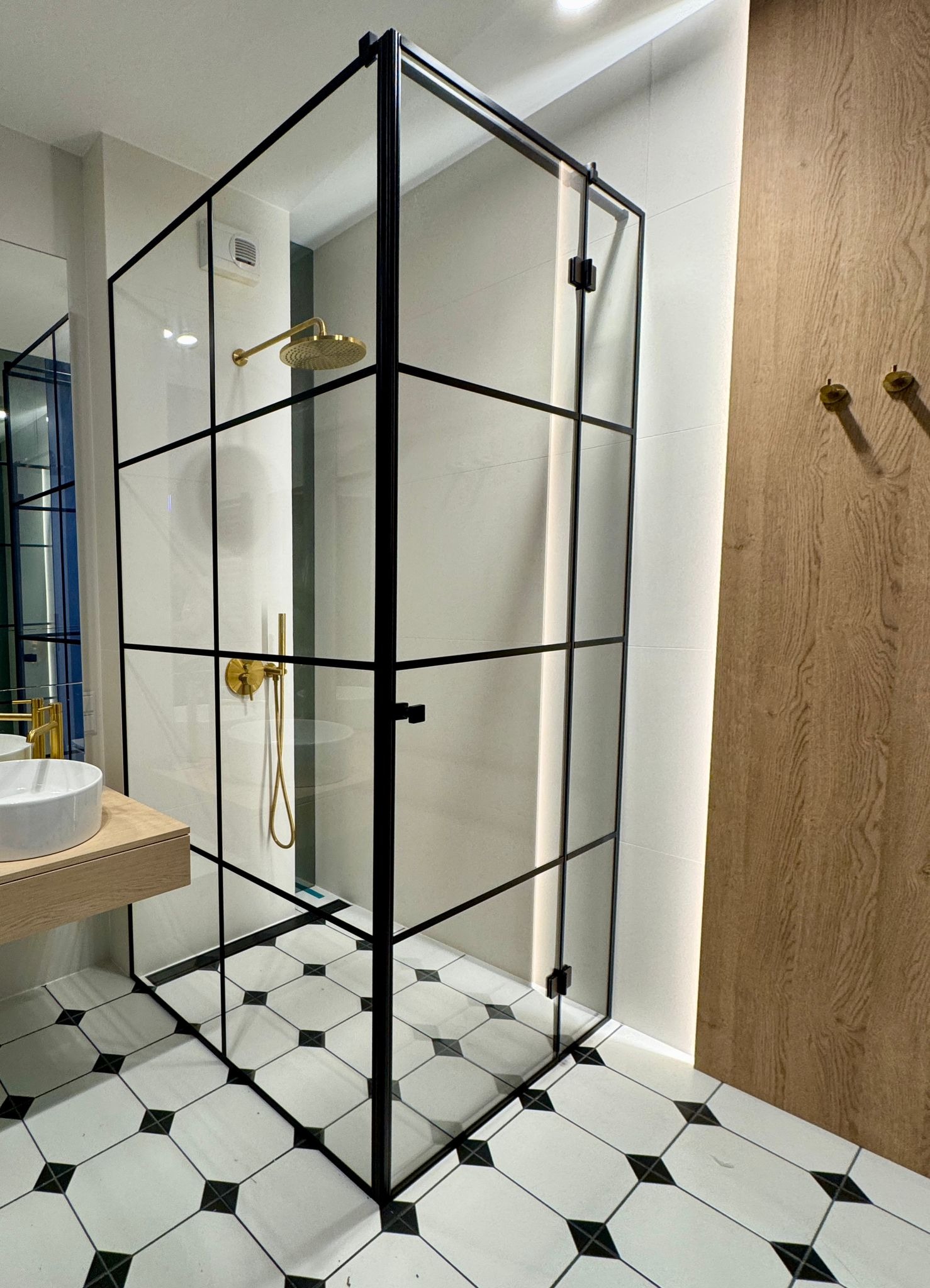 Nowoczesna kabina prysznicowa ze szklanymi ścianami z czarnymi profilami, złota armatura, biało-czarne płytki podłogowe w łazience.