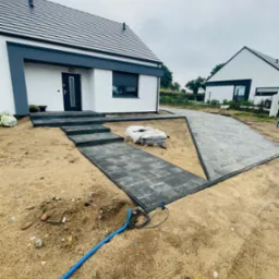 Nowoczesny dom z szarą kostką brukową prowadzącą do wejścia i podjazdu, widoczne worki z materiałem i niebieski wąż na piasku.