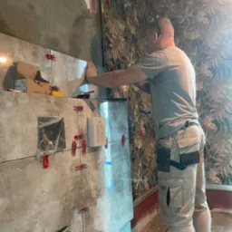 Mężczyzna w szortach i okularach montuje szare płytki na ścianie, używając czerwonych klipsów poziomujących, widoczna paczka płytek Stalco i instalacja podtynkowa Grohe.