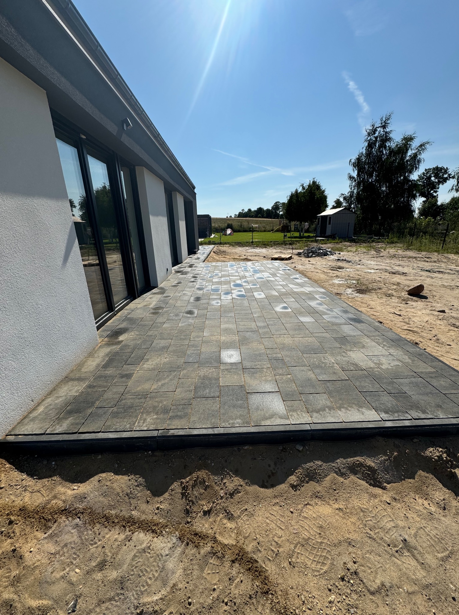 Nowo ułożona szara kostka brukowa z delikatnym wzorem, przylegająca do ściany budynku z dużymi oknami, widoczne ślady na piasku i fragment ogrodu w tle.