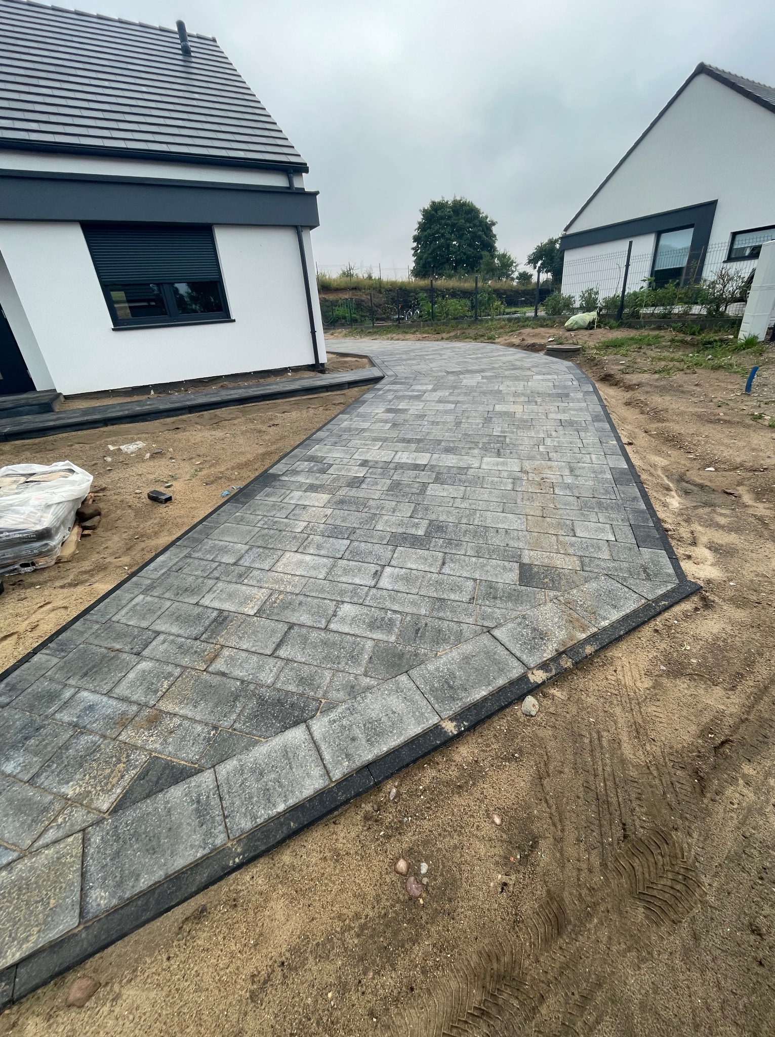 Ułożona ścieżka z szarej kostki brukowej prowadząca do nowoczesnego domu z białymi ścianami i czarnymi akcentami, widoczne ślady opon na piasku po bokach.