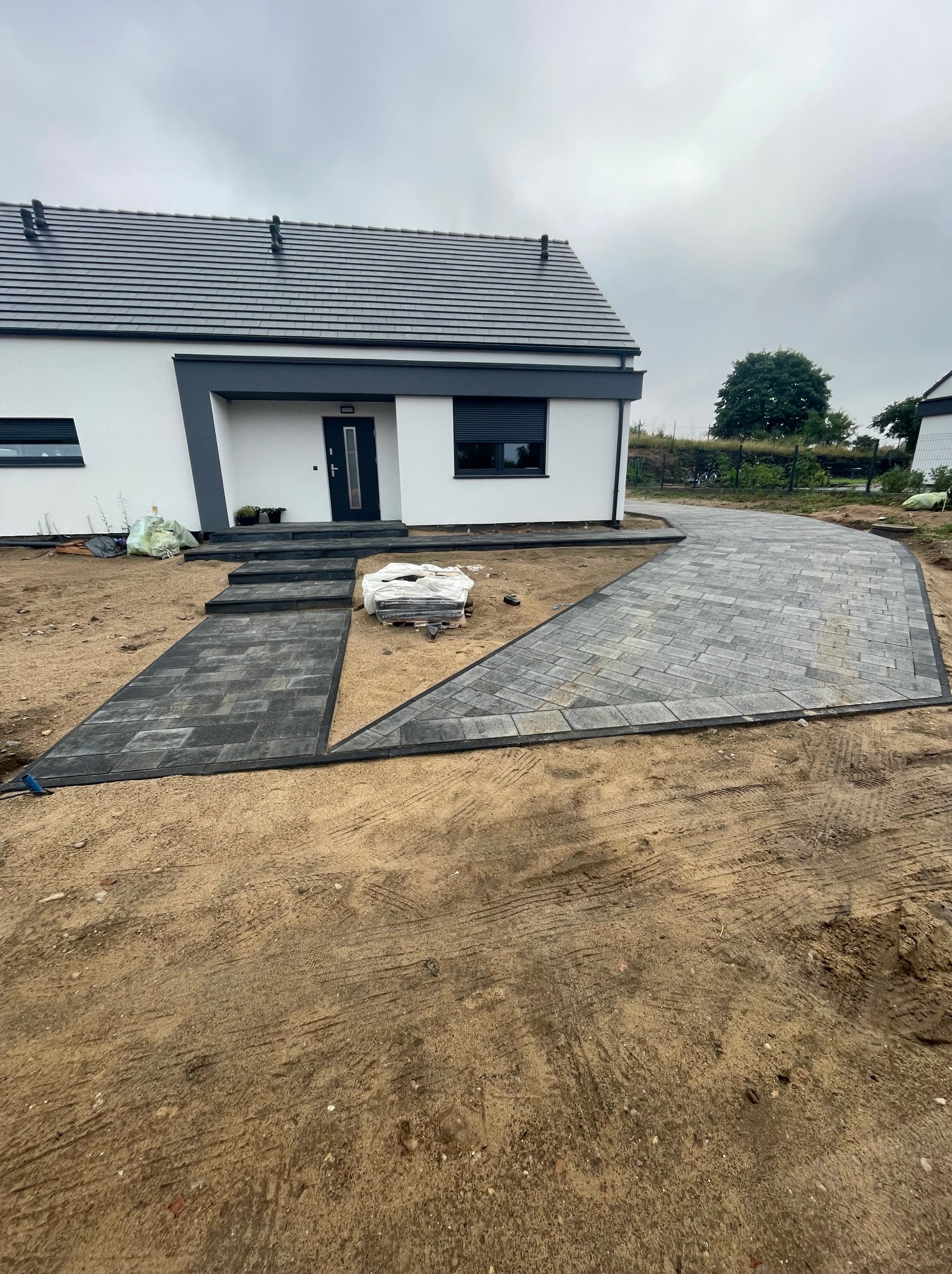 Nowoczesny dom z antracytową kostką brukową ułożoną w geometryczny wzór, widoczne schody wejściowe i fragment podjazdu, w tle trawnik i ogrodzenie.