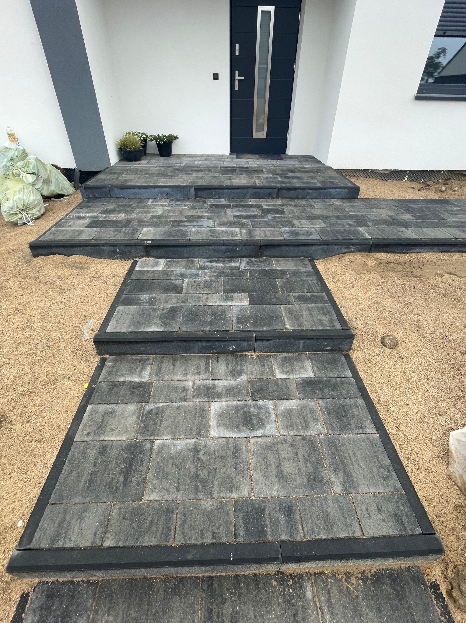 Nowoczesne schody wejściowe i ścieżka z szarej kostki brukowej z czarnym obrzeżem, prowadzące do białego domu z ciemnymi drzwiami, otoczone piaskiem i workami z materiałami budowlanymi.