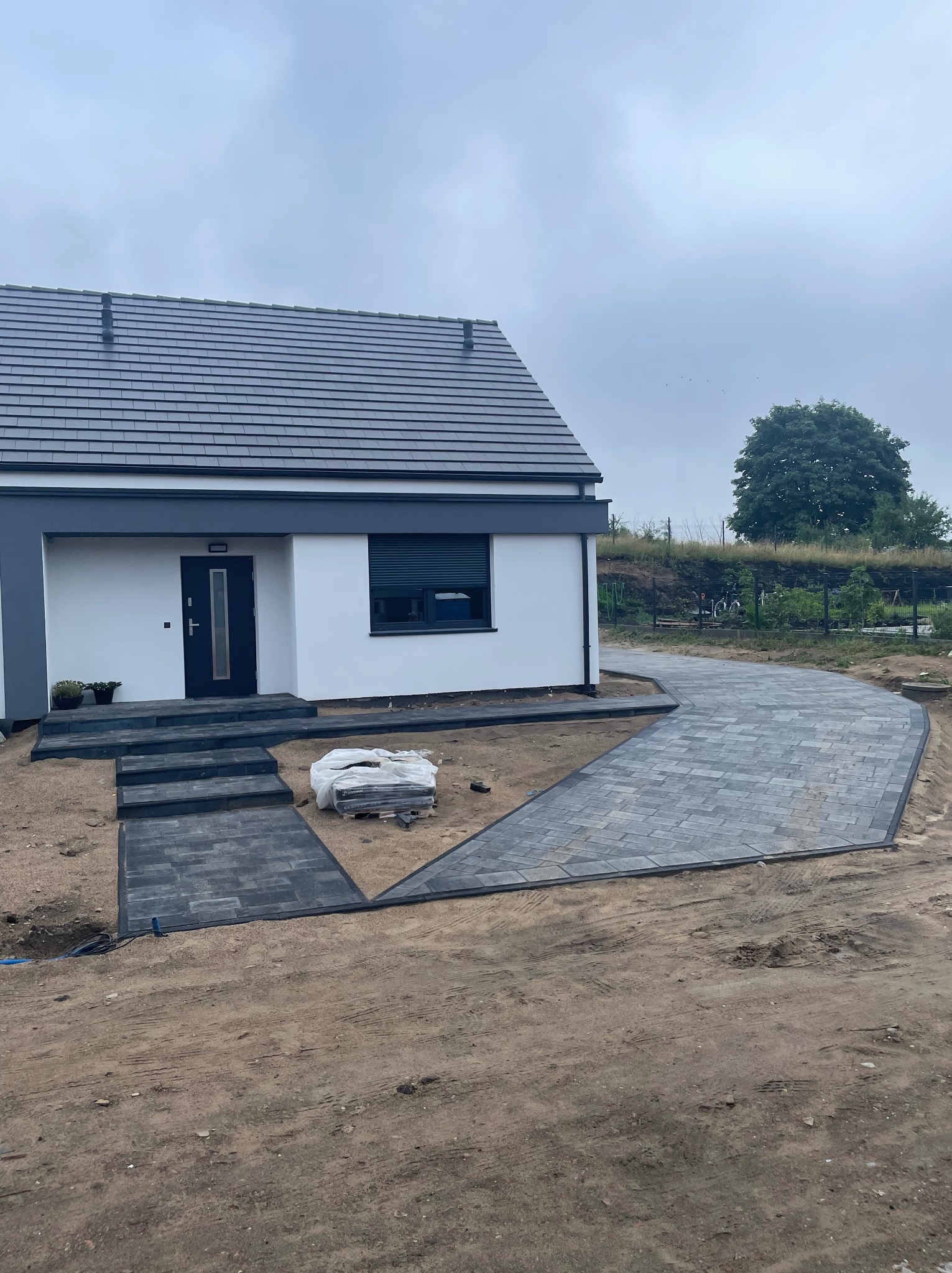 Ułożona z szarej kostki brukowej ścieżka i schody prowadzące do nowoczesnego domu z ciemnymi oknami i dachem; paleta kostki brukowej obok schodów.