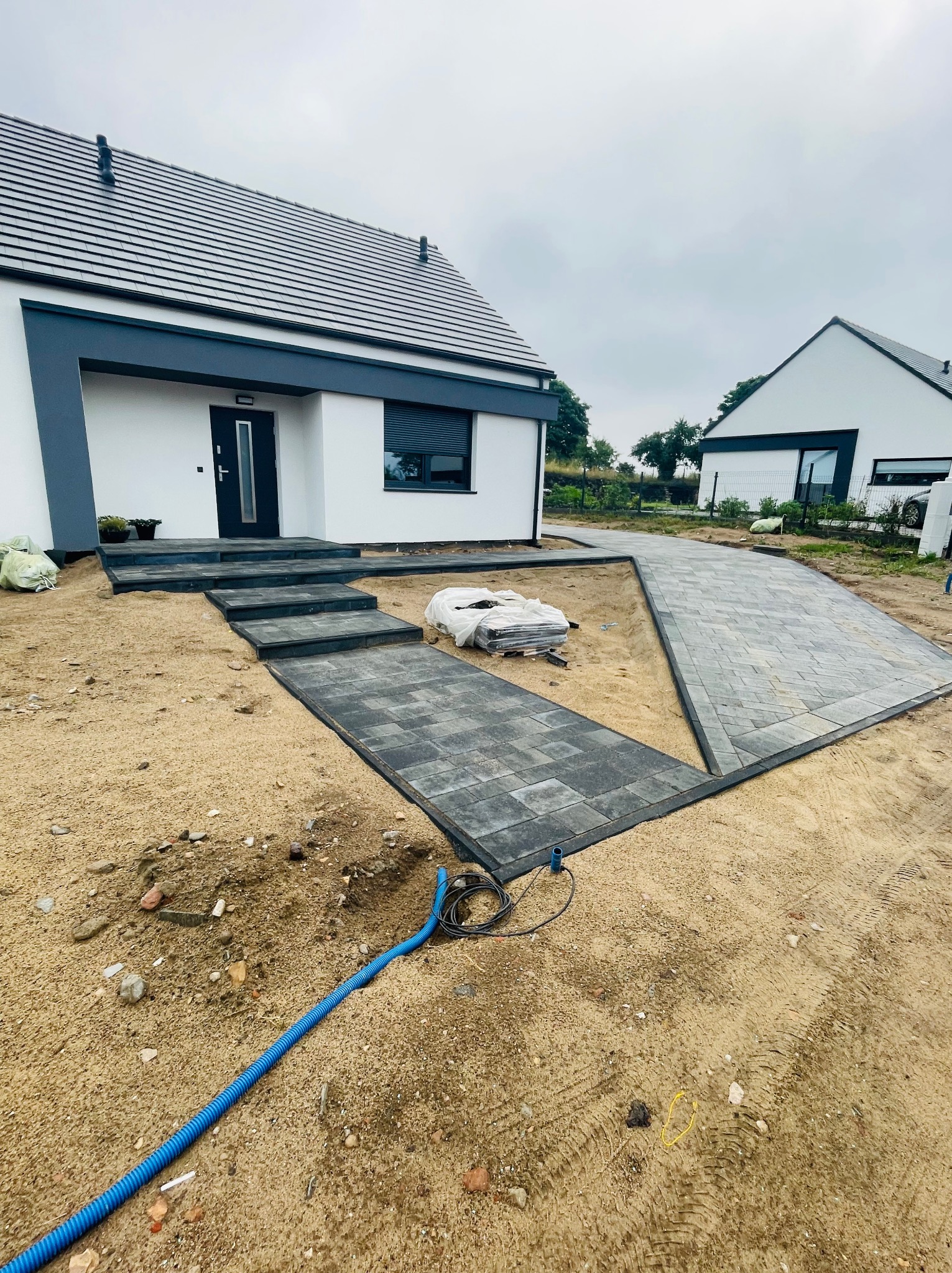 Nowoczesny dom z szarą kostką brukową prowadzącą do wejścia i podjazdu, widoczne worki z materiałem i niebieski wąż na piasku.