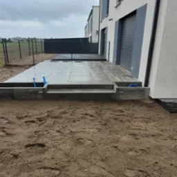 Nowoczesny taras z szarych płyt betonowych, z niską donicą i schodami, widoczne rury instalacji wodnej, w tle ogrodzenie panelowe i budynki mieszkalne.