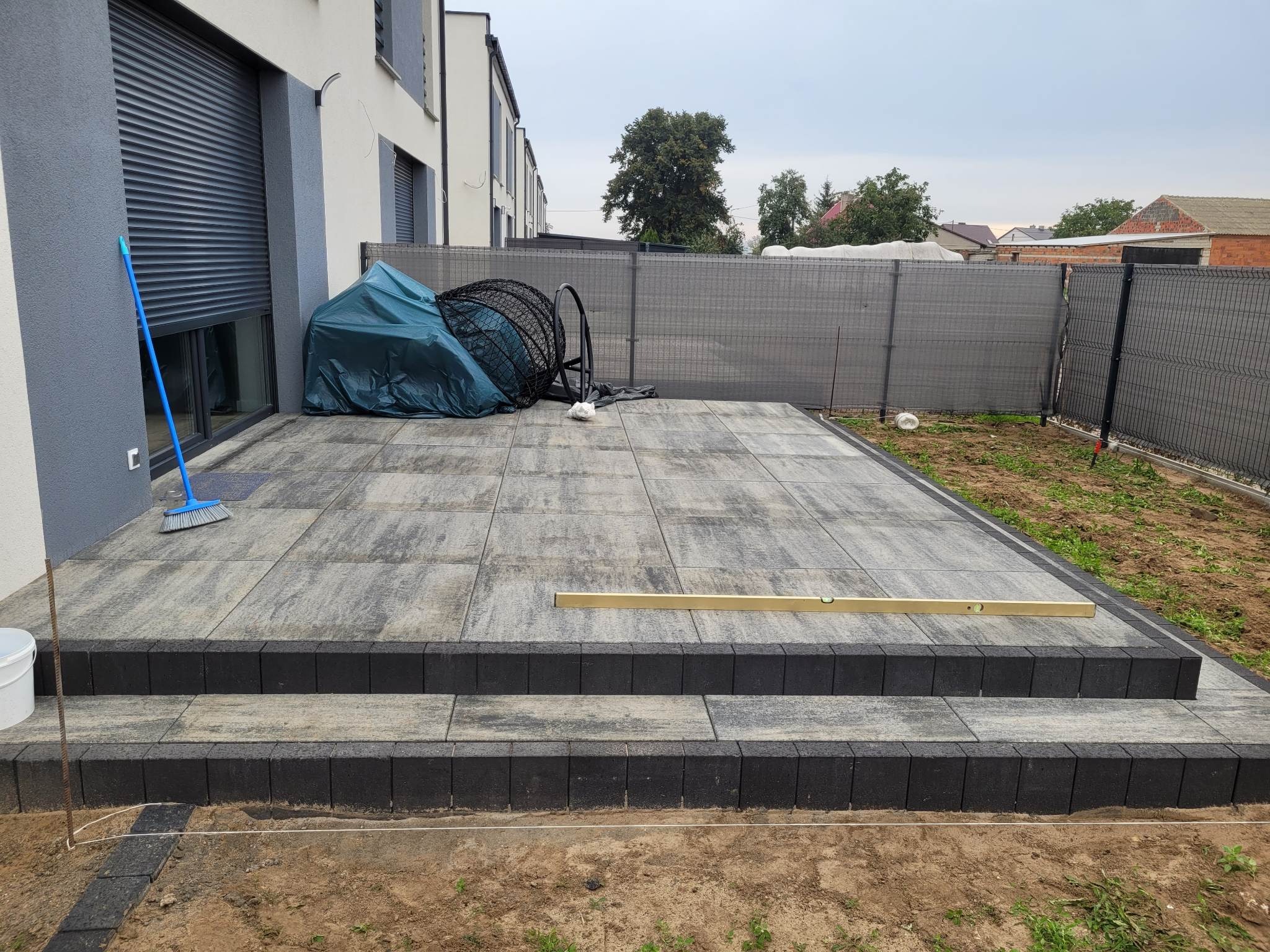 Nowo ułożony taras z szarej kostki brukowej z czarnymi obrzeżami i schodami, poziomica leży na jednym ze stopni, w tle siatka ogrodzeniowa i budynek.
